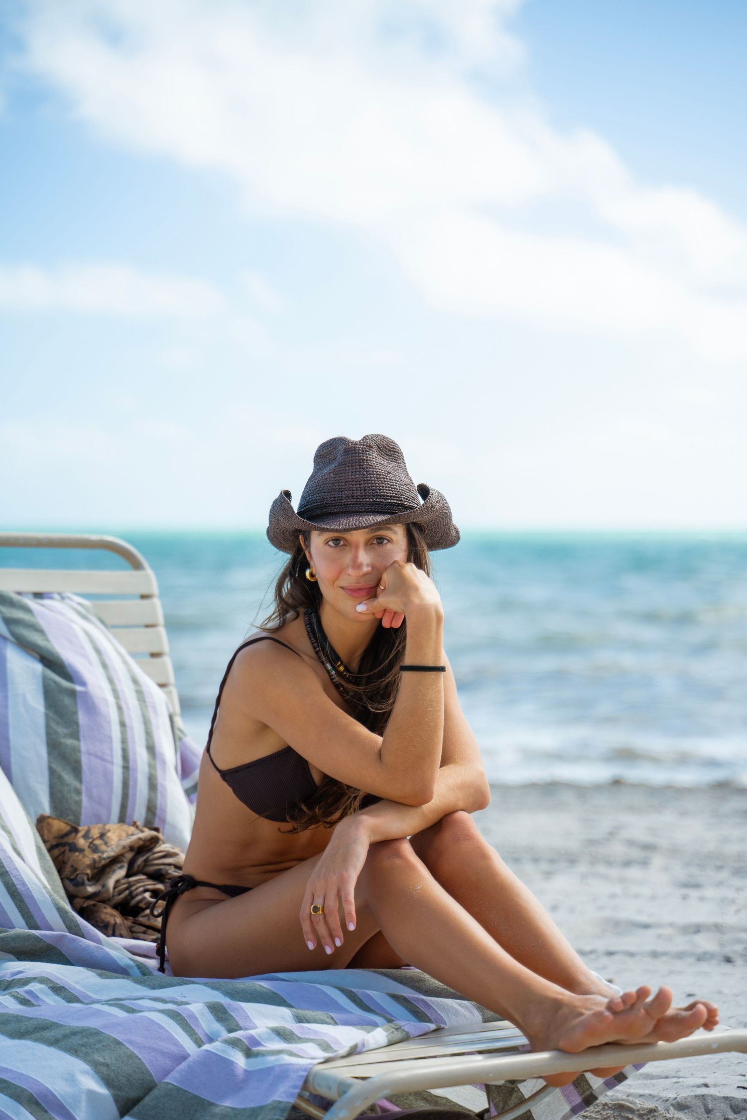 Woman in black swimsuit and hat relaxes on striped beach chair by turquoise ocean waters.