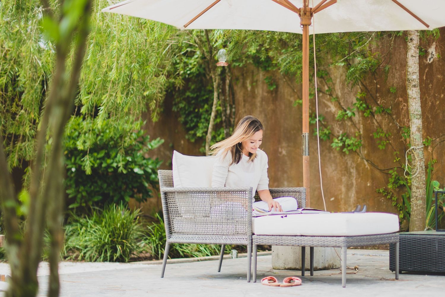 Someone relaxes on a white outdoor lounge chair under a patio umbrella in a lush garden setting.