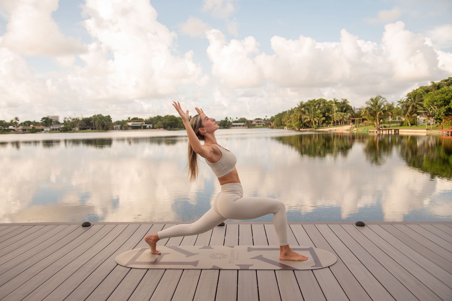 Yoga flow demonstration on a lakeside pier with palm trees and cloudy sky in the background.
