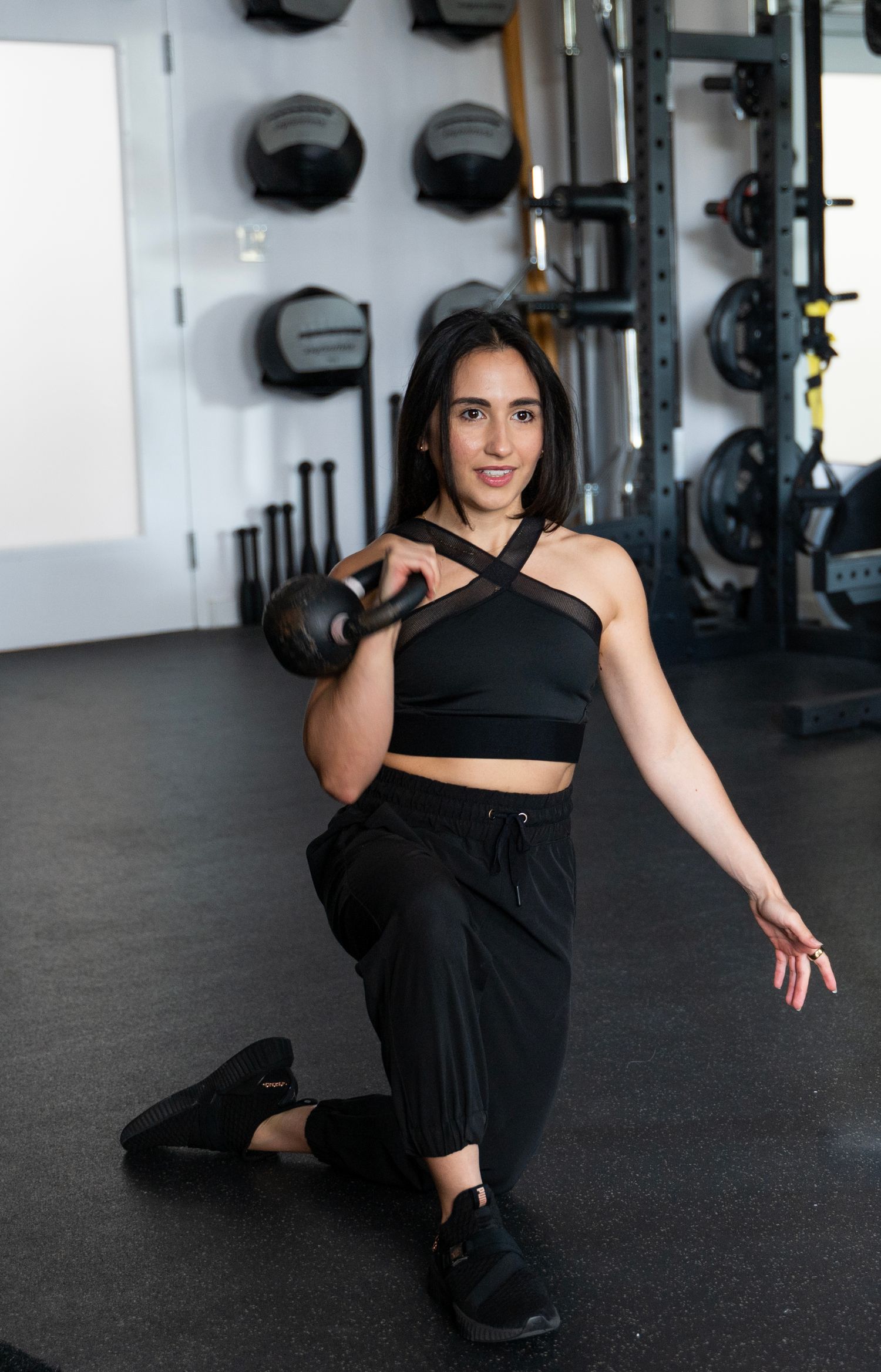 Woman in black athletic wear poses on gym floor with exercise equipment visible in background.
