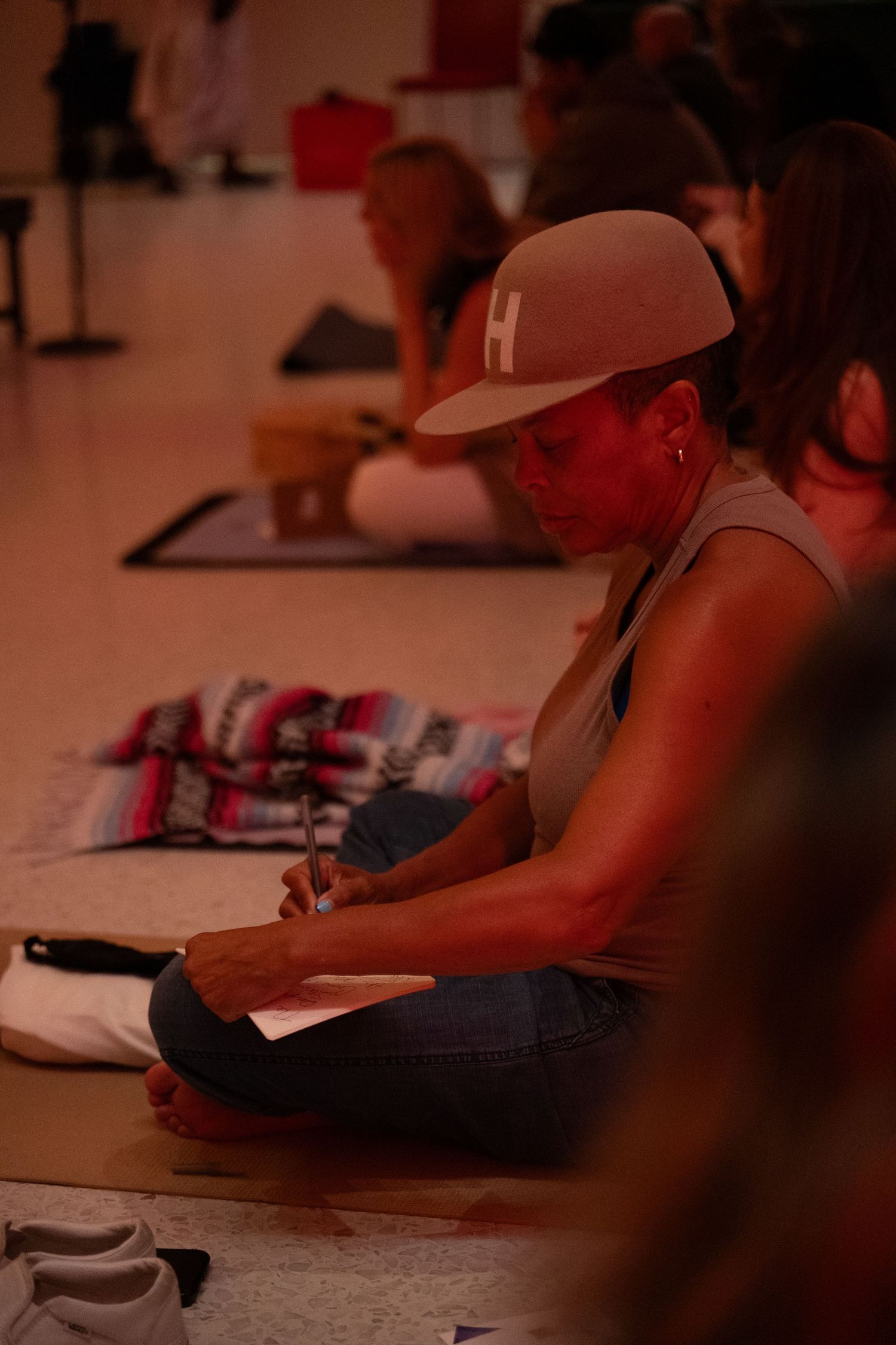 Woman in gray cap sits cross-legged on yoga mat, writing in notebook during a group wellness event indoors.