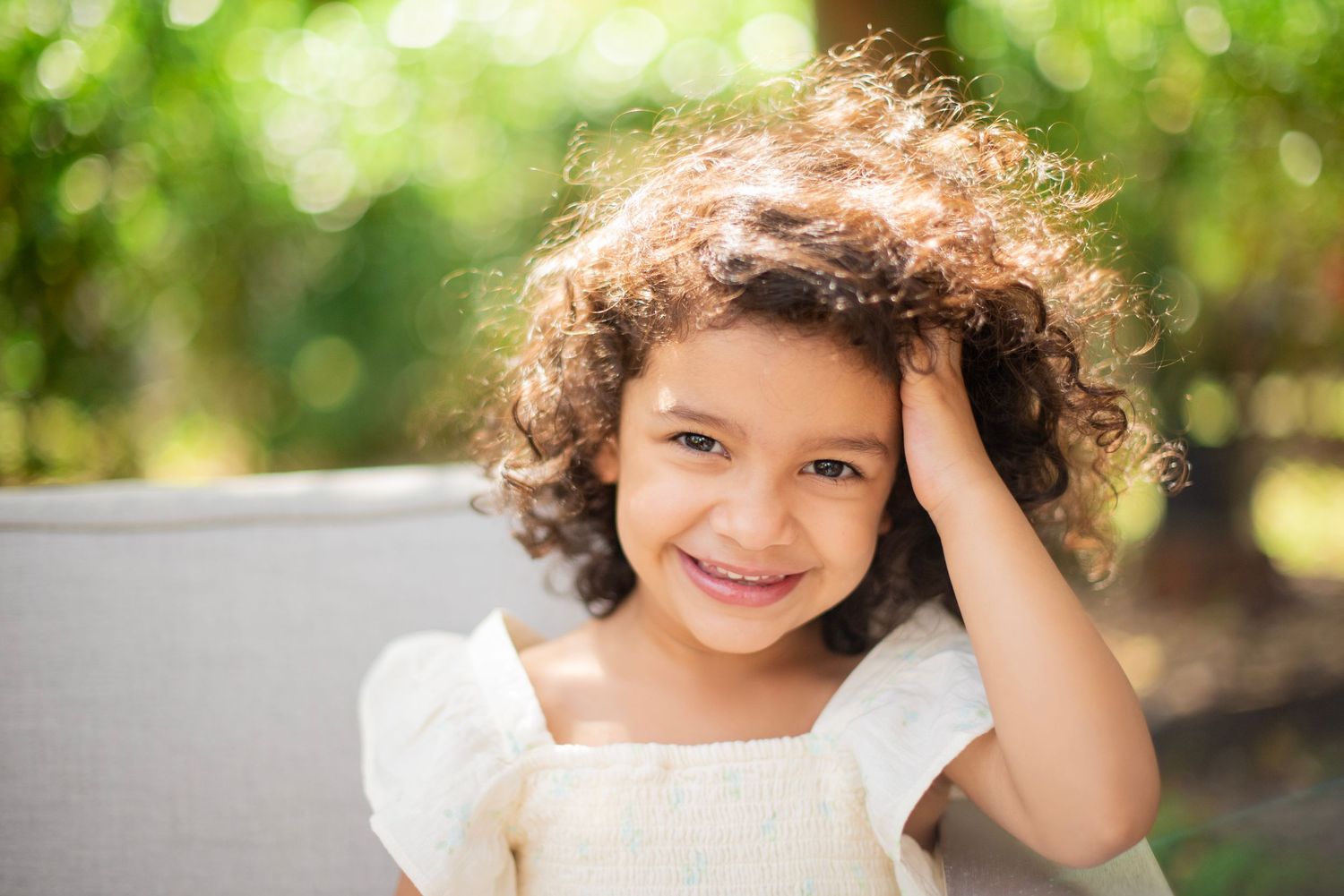 A child with curly hair poses outdoors in natural lighting against a blurred green background.