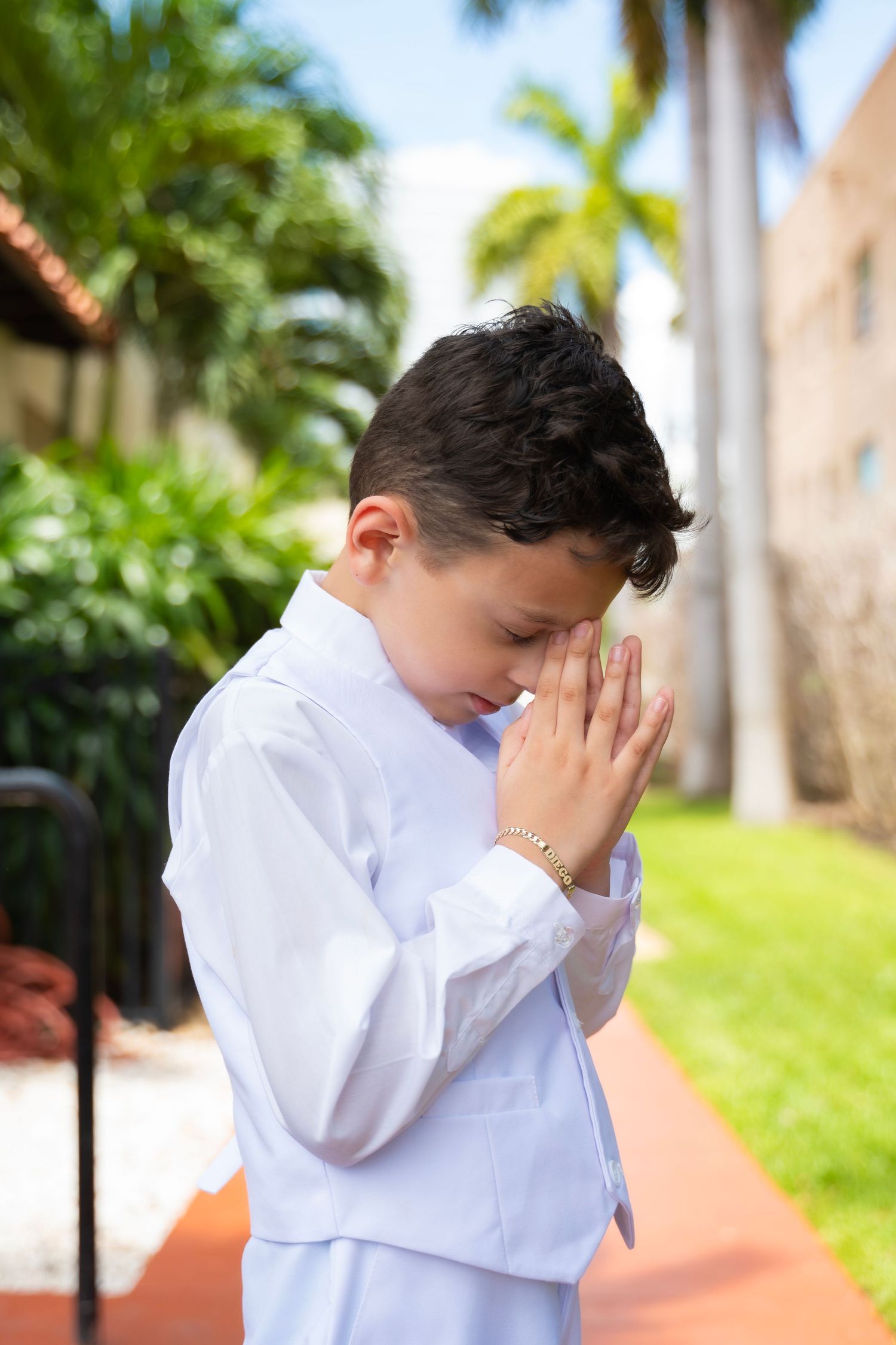 Young boy in white suit praying outdoors at his First Communion ceremony.
