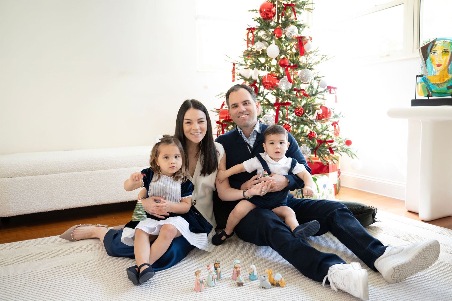 Family lounging on floor near decorated Christmas tree in white living room during holiday season.