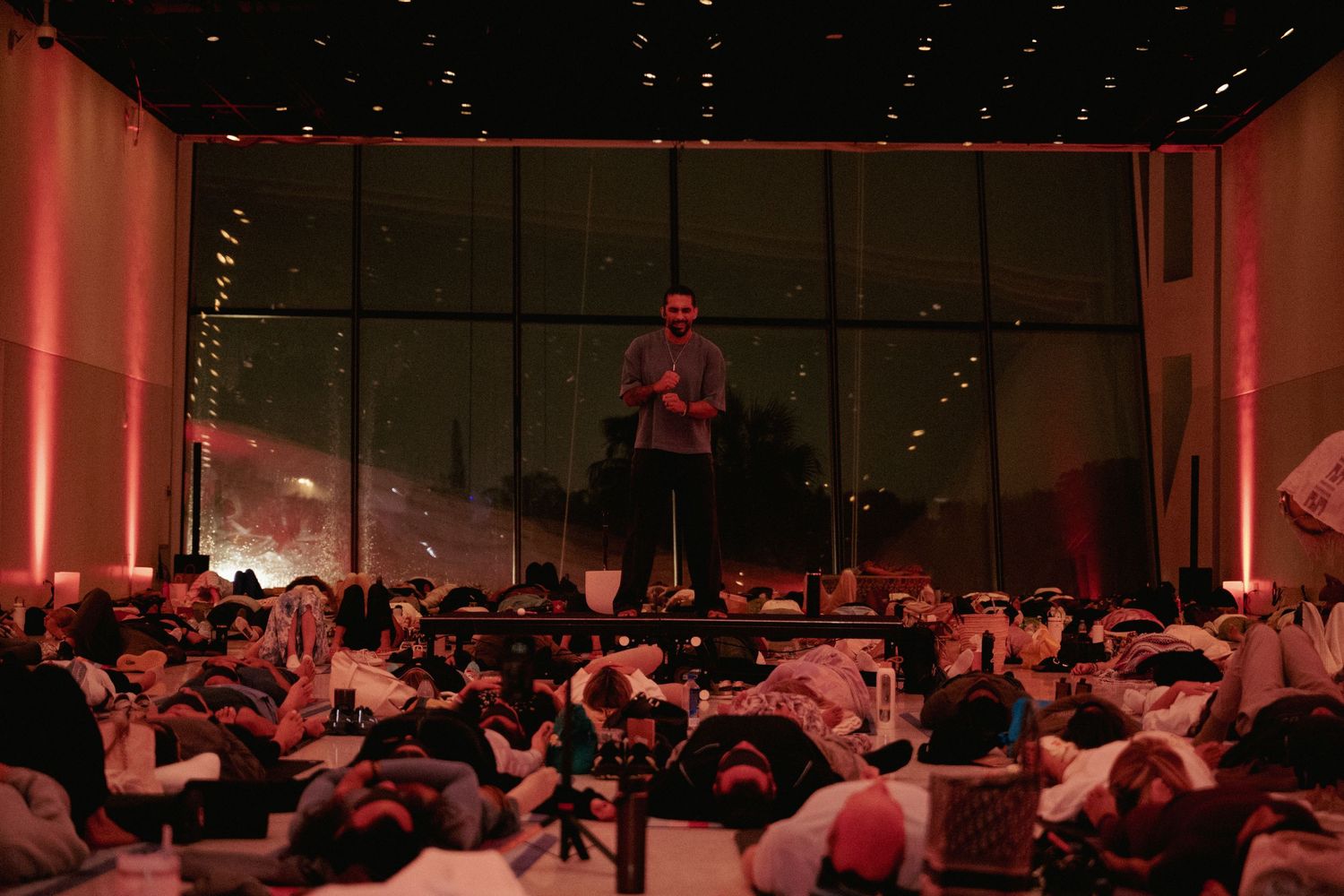 A speaker stands on stage addressing a large audience lying on the floor in a dimly lit red venue.