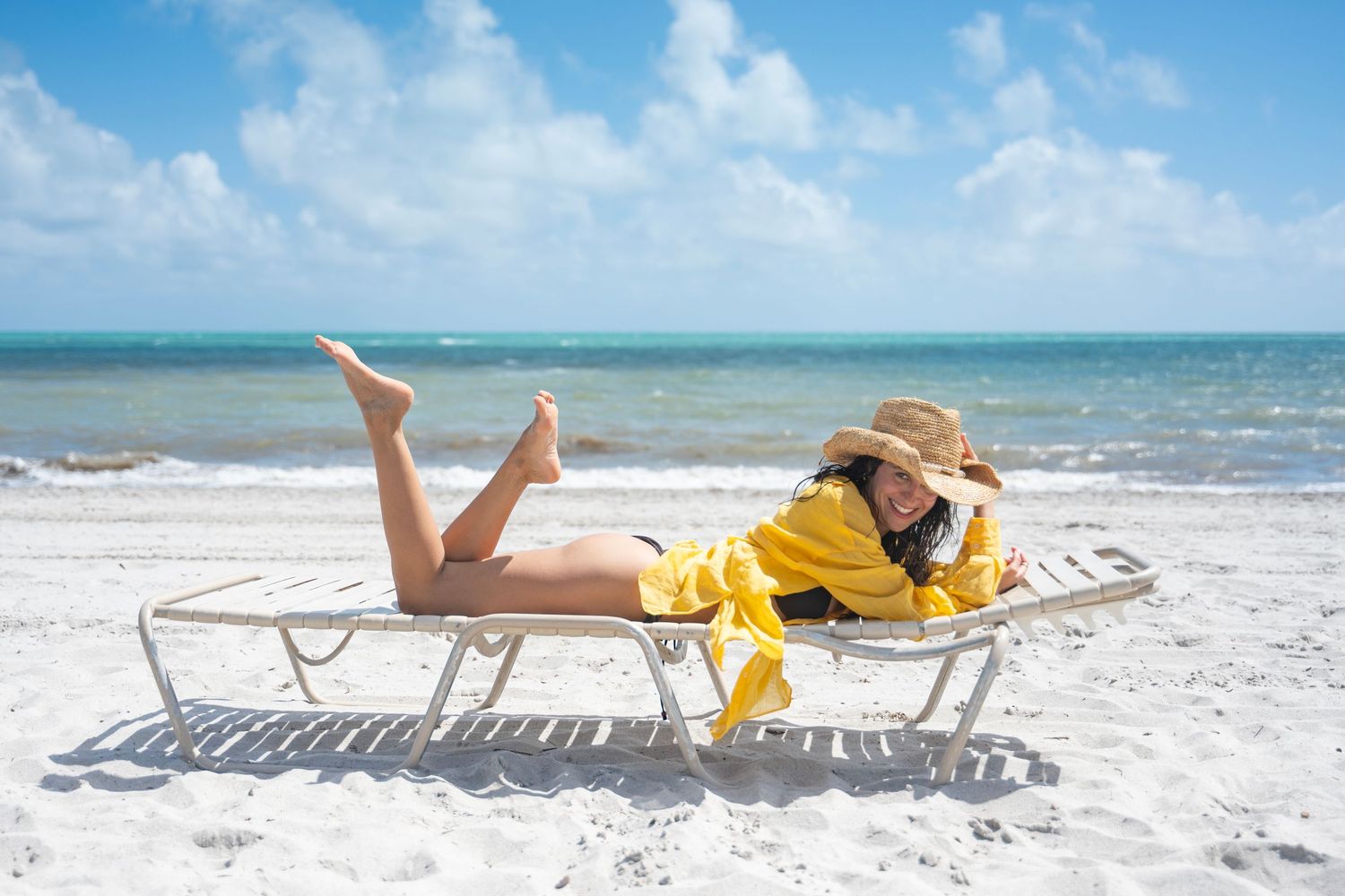 Person relaxing on a white lounge chair at a sunny beach with turquoise ocean waves in the background.