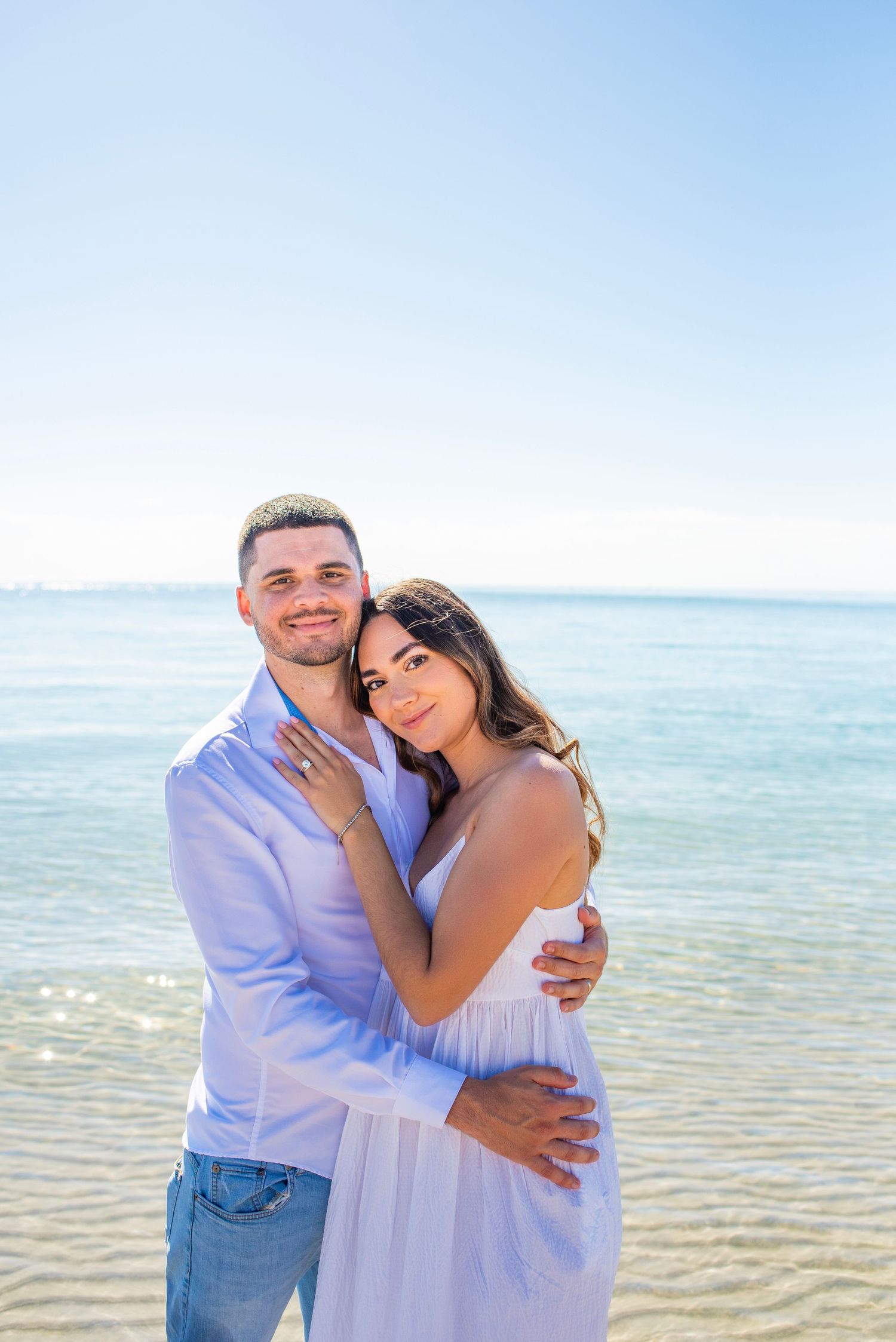 A couple embraces lovingly on a sunny beach with crystal clear waters and blue sky in the background.