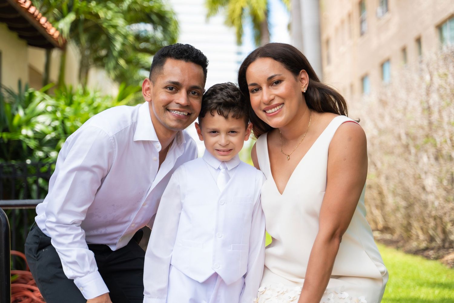 Family of three smiling outdoors, parents in white attire with young boy in white suit, lush green background.