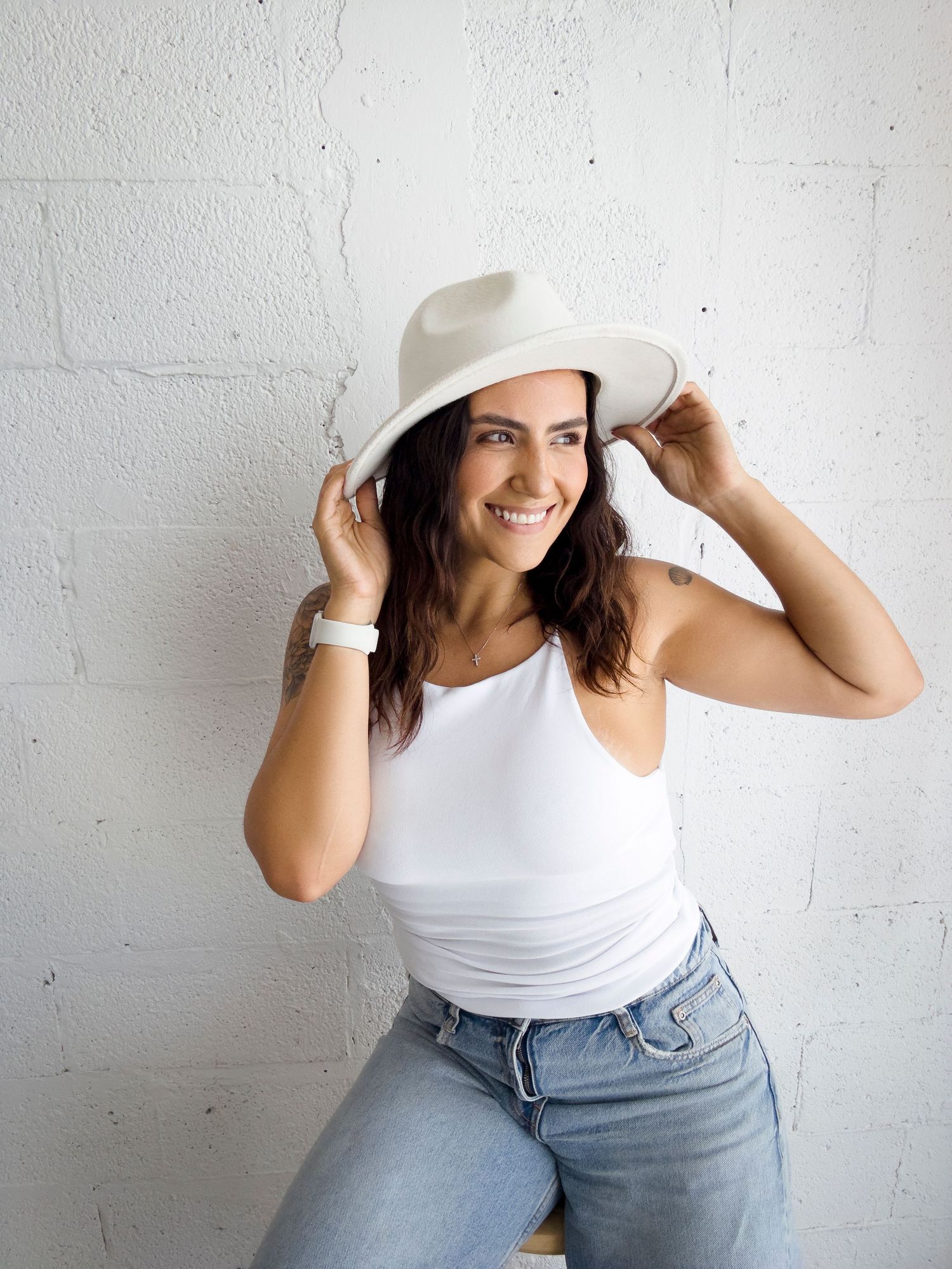 Candid shot of woman adjusting white hat while posing against white wall.
