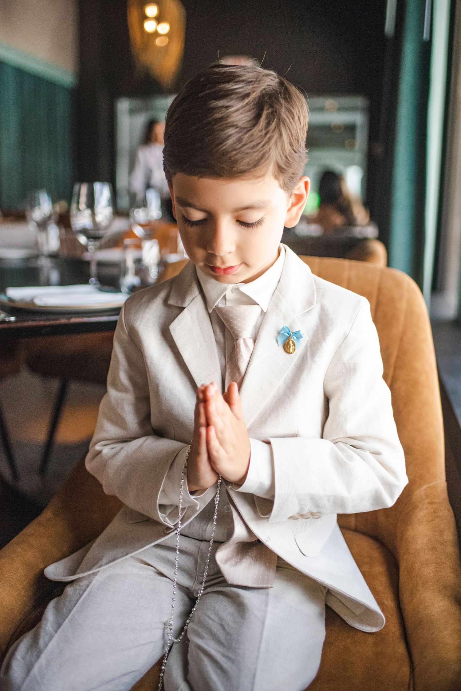 Child in formal white attire sitting thoughtfully on brown leather couch.