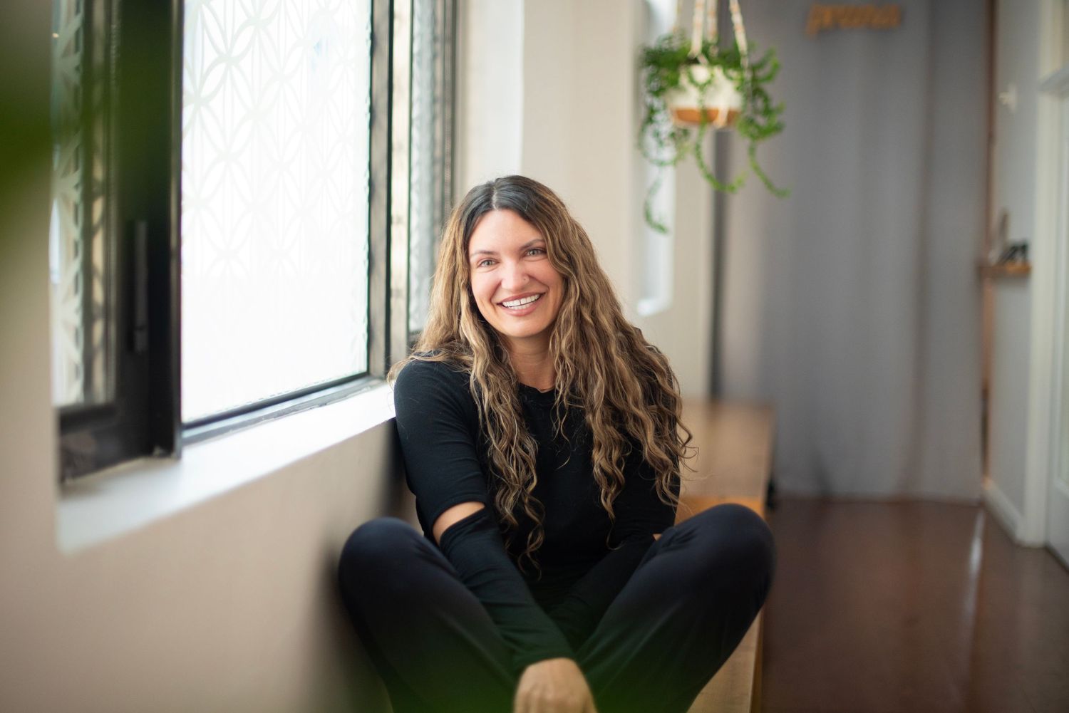 Someone sits cross-legged on hardwood floor near window in bright interior space with hanging plant decor.