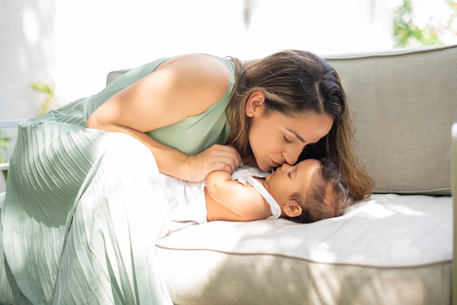 A mother and baby share a tender moment while lying together on a couch in a sunlit room.