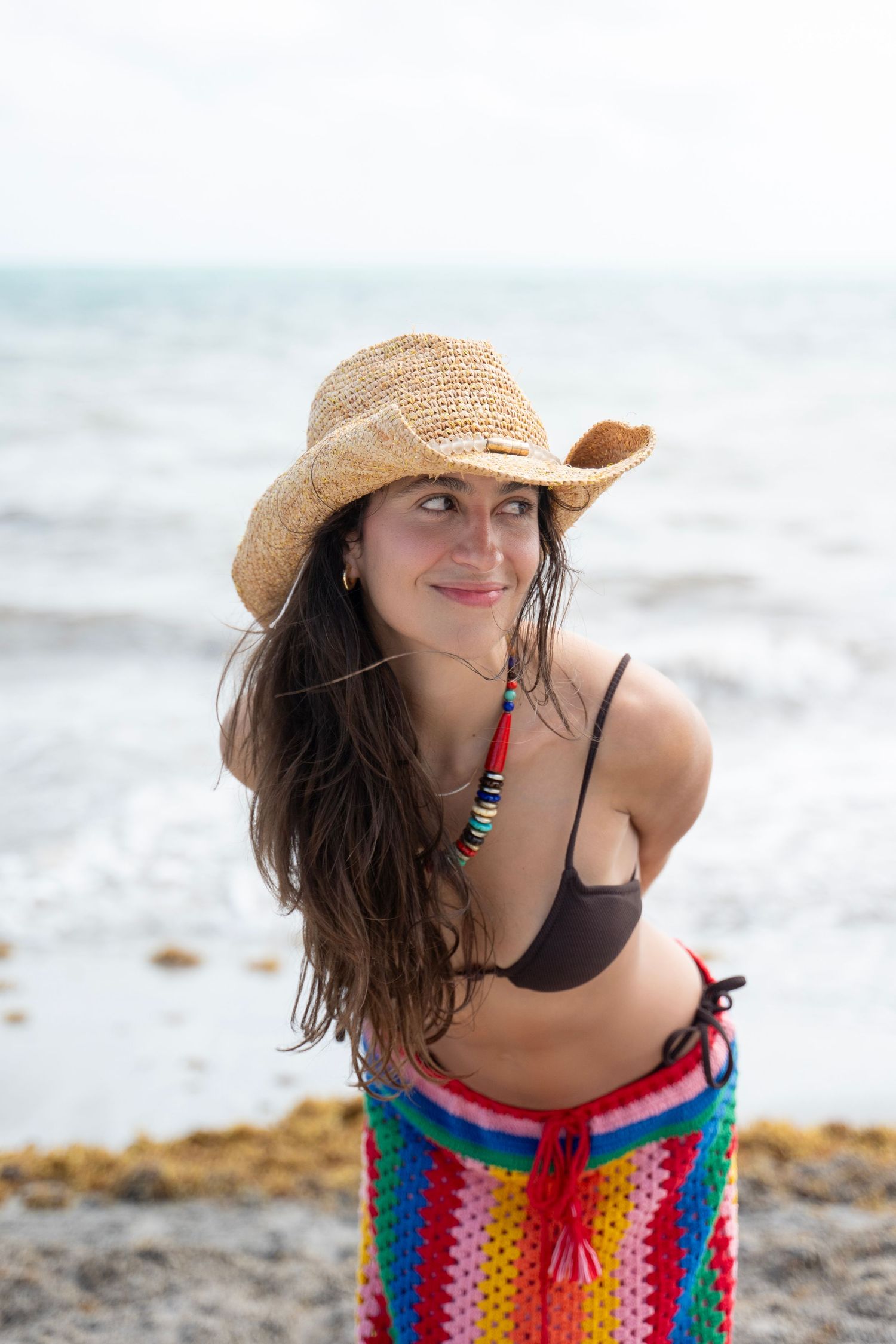 A person wearing a colorful striped cover-up and straw hat poses playfully on a beach.