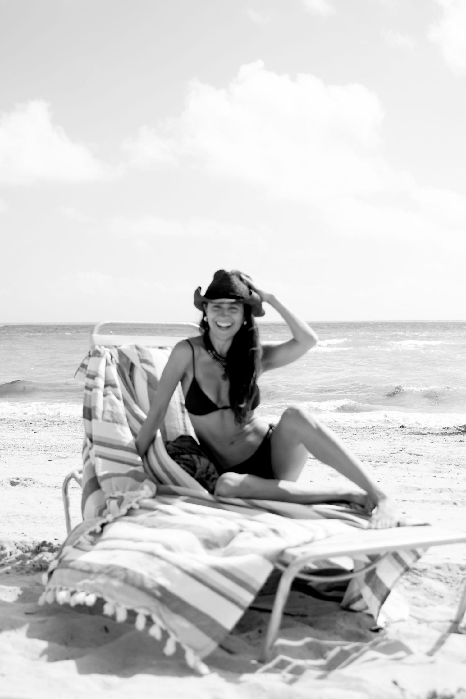 Black and white photo of someone relaxing on a striped beach blanket by the ocean wearing a bikini and sun hat.
