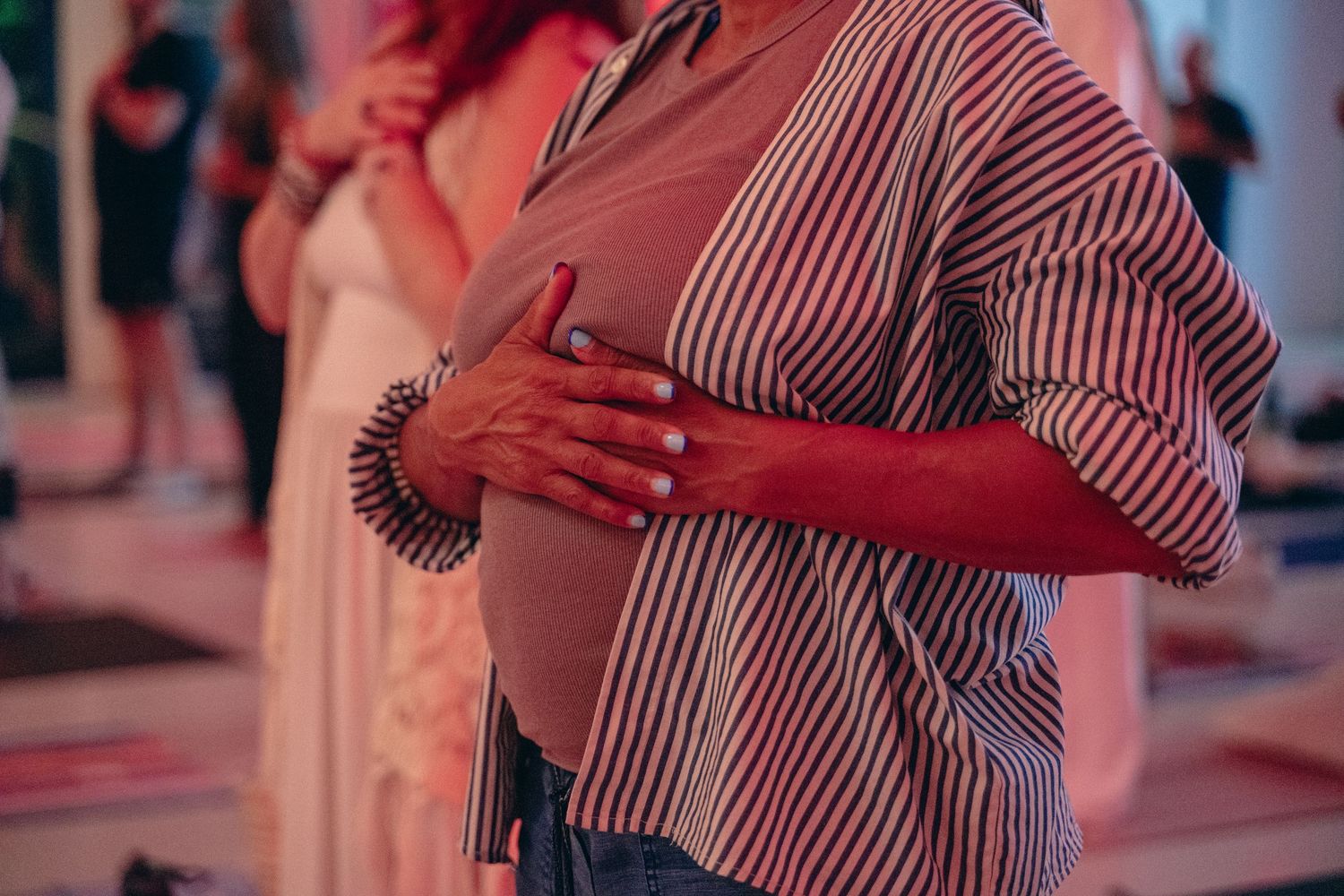 A pregnant woman in a striped shirt gently holds her baby bump at an indoor event with pink lighting.