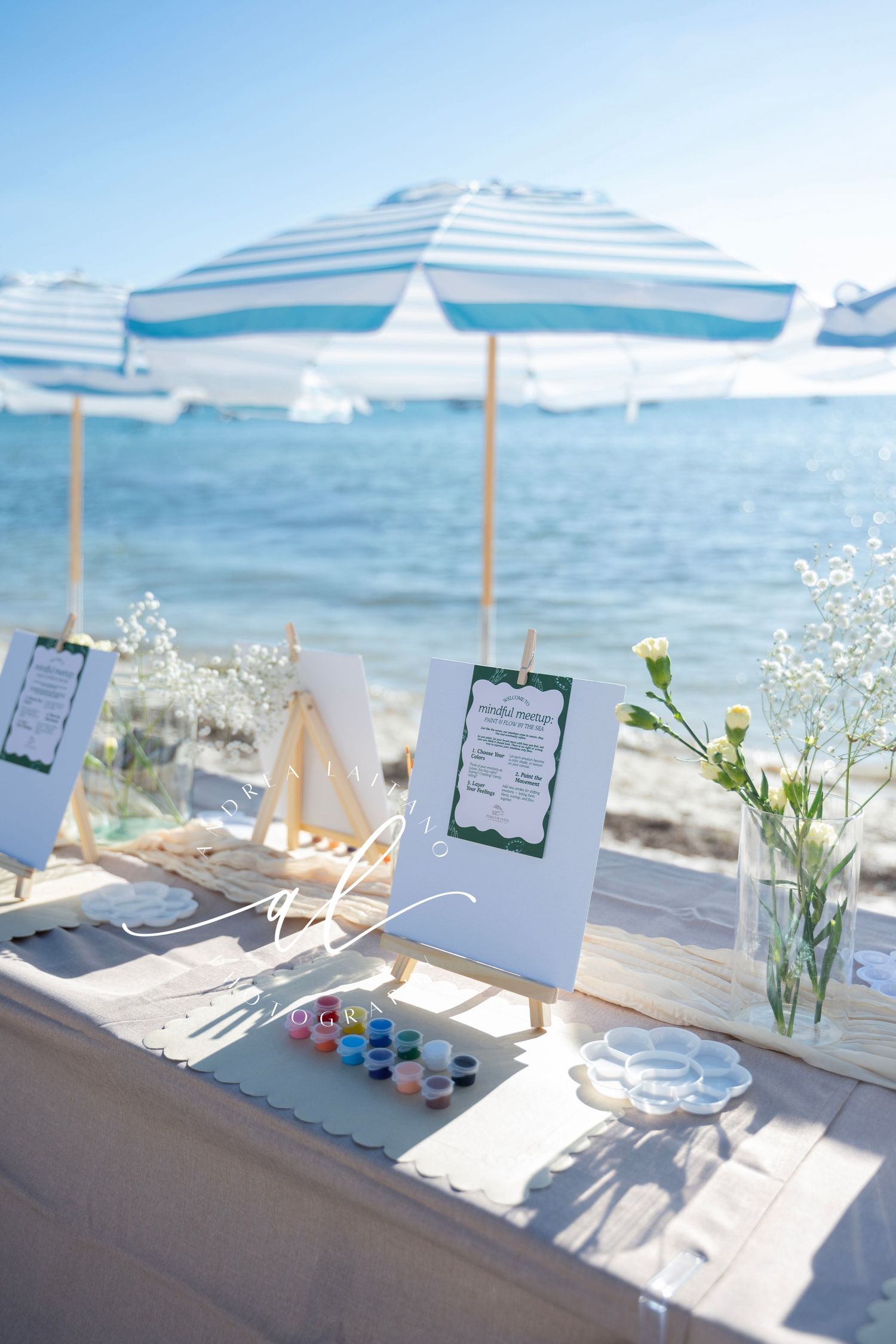 Beachside table setup with blue striped umbrellas, clipboards, and white floral arrangements against an ocean backdrop.