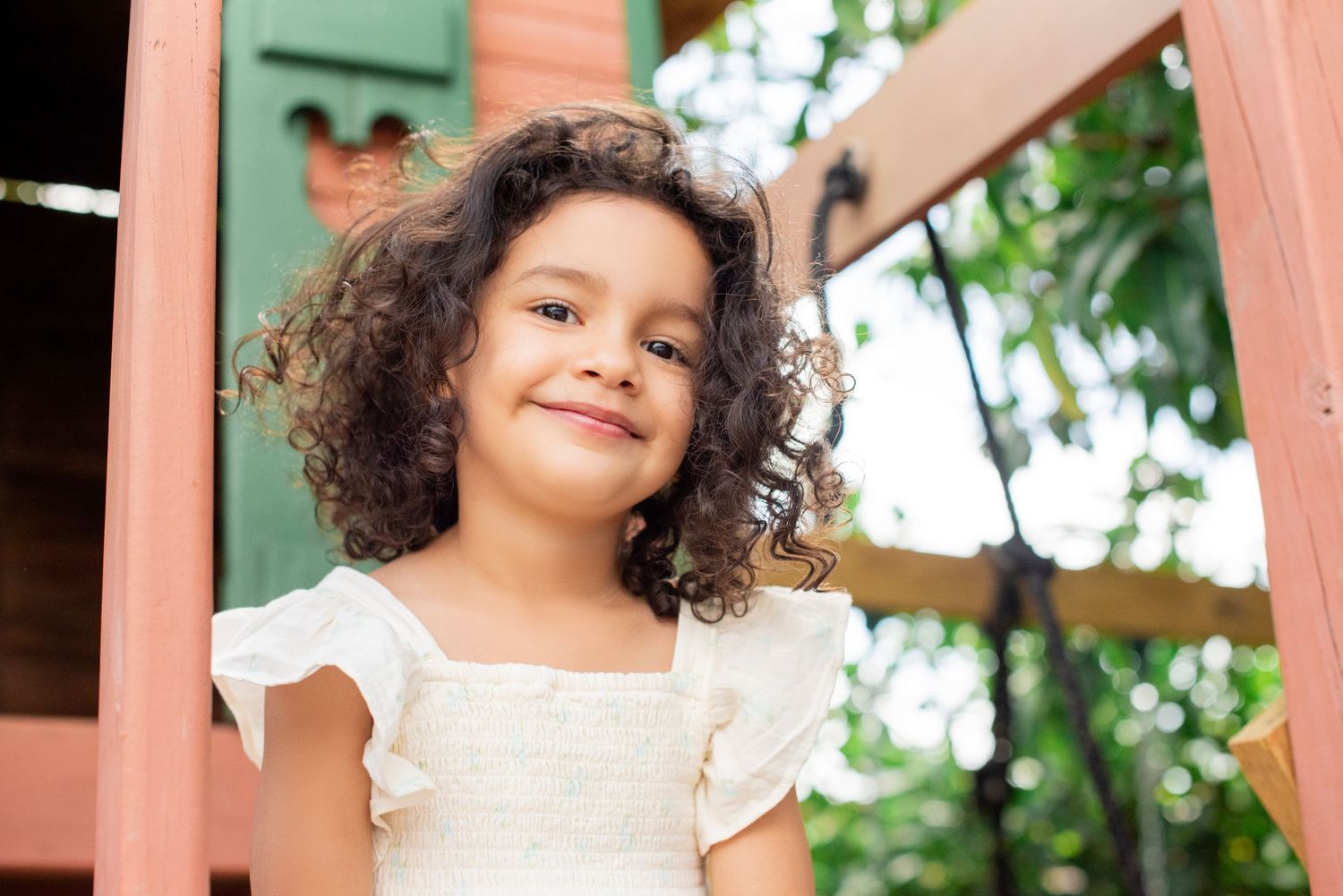 A young child with curly hair smiles while playing outdoors near pink-tinted wooden posts.