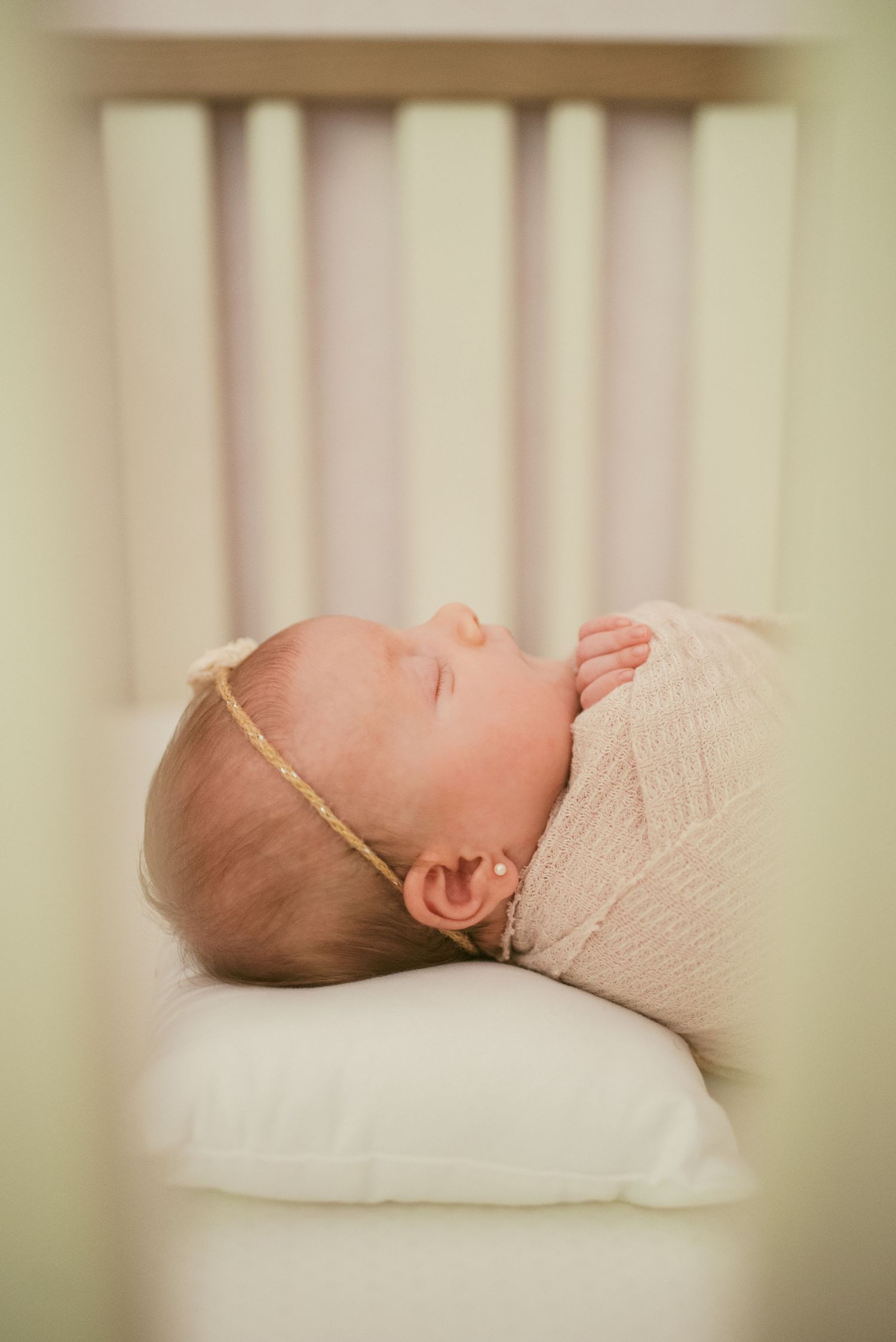 Sleeping newborn baby wearing white knit outfit rests peacefully on white pillow in soft natural lighting.