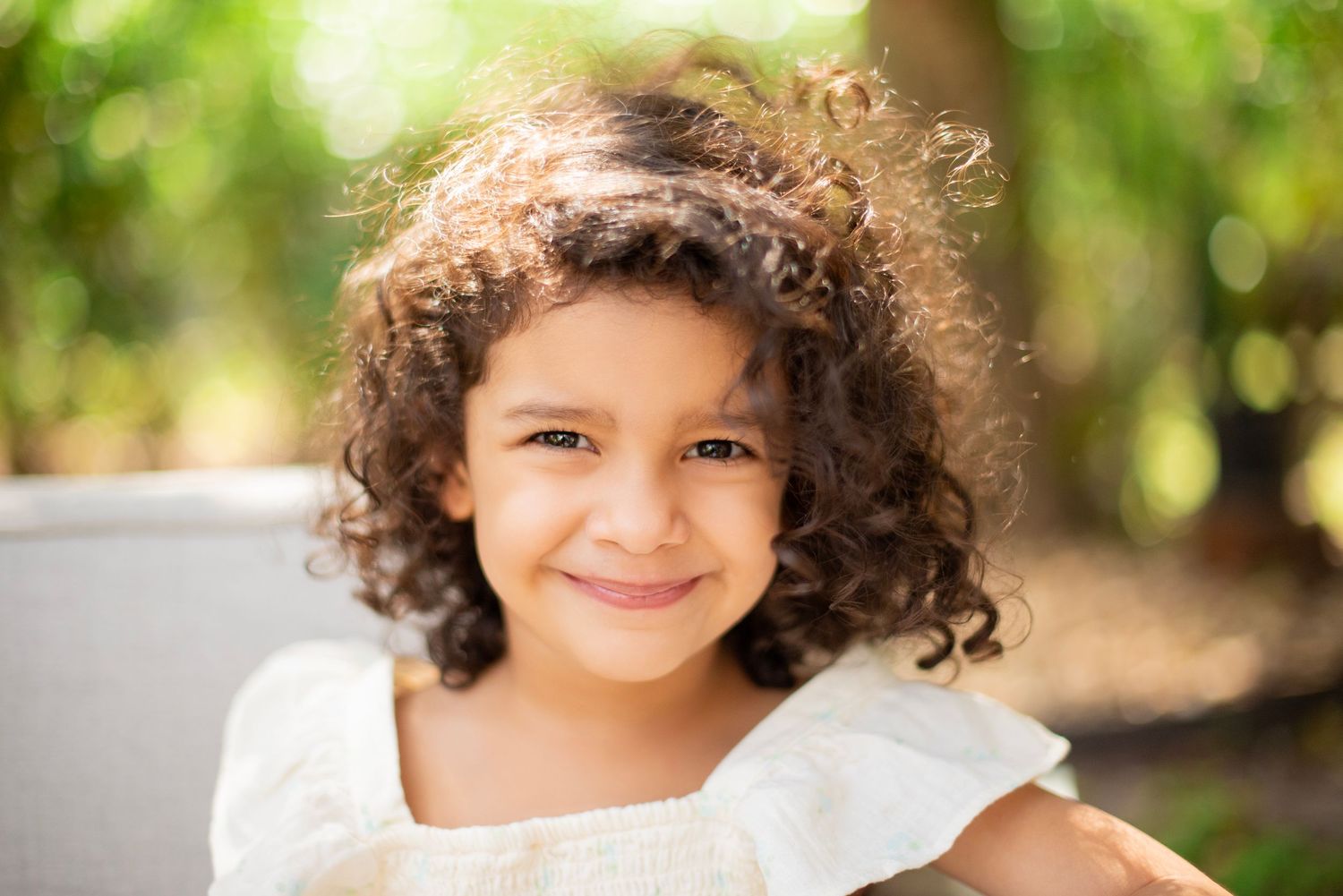 Child with curly hair smiling in outdoor portrait series with natural lighting and greenery in background.