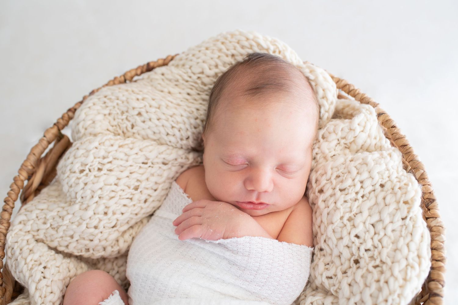 Sleeping newborn wrapped in white fabric on knitted cream blanket.