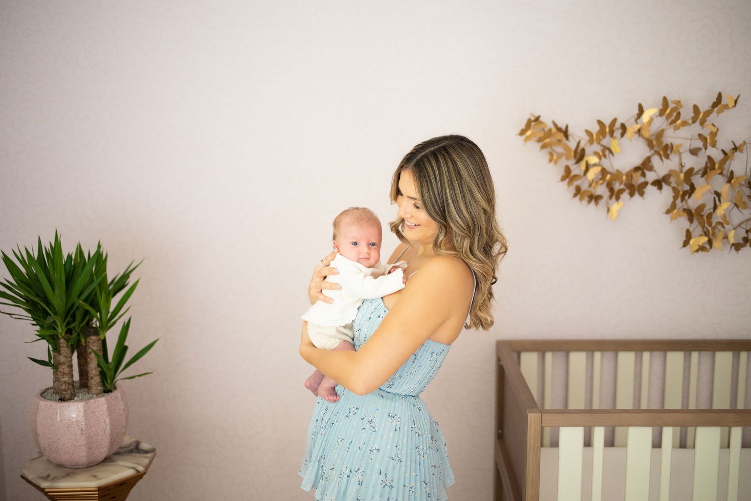 Parent in blue dress holds and comforts baby in white nursery with natural decor and wooden accents.