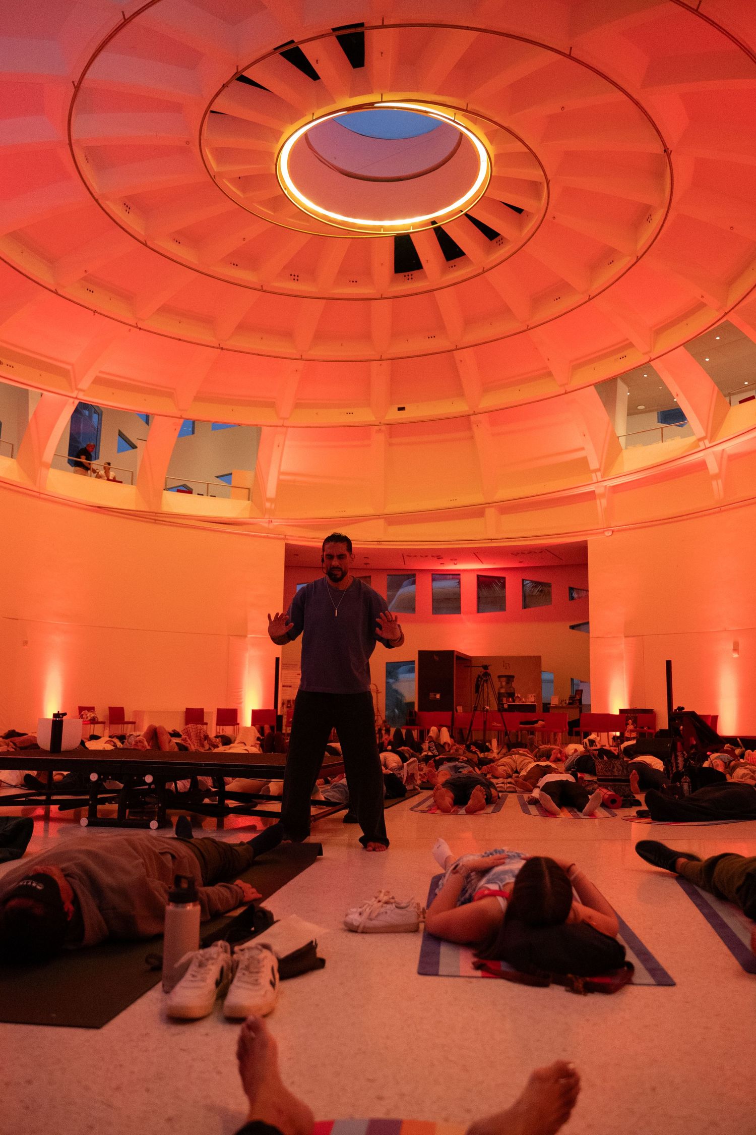 Yoga class in a domed rotunda with warm red lighting, participants lying on mats while instructor stands center.