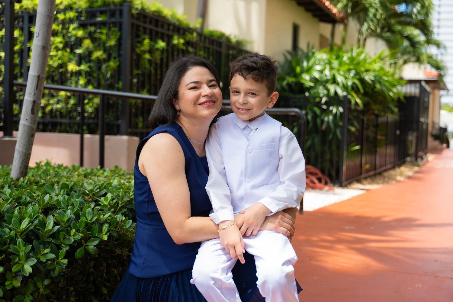 A smiling woman in a navy dress holds a young boy in white clothes outdoors near lush greenery.