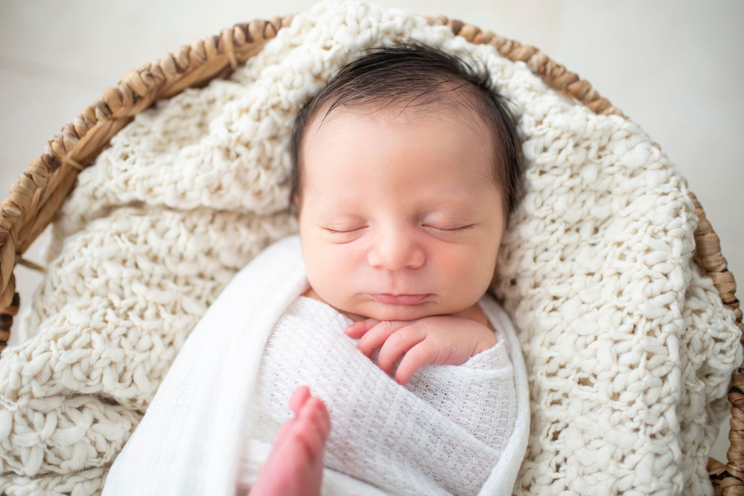 Sleeping baby nestled in round basket with white textured blanket.