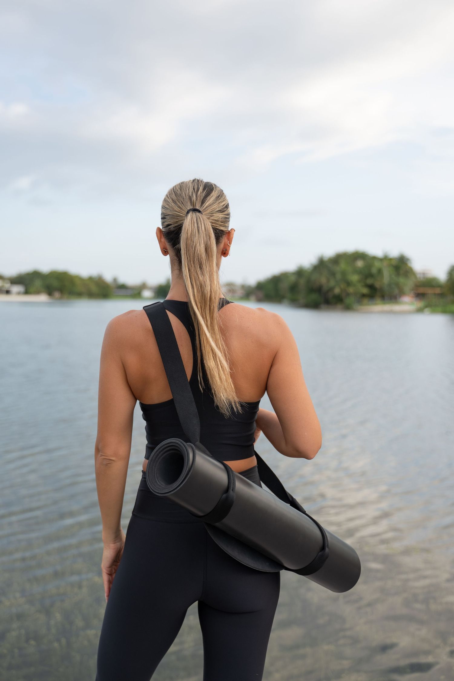 Athletic woman in black workout attire carrying a yoga mat by a scenic waterfront.