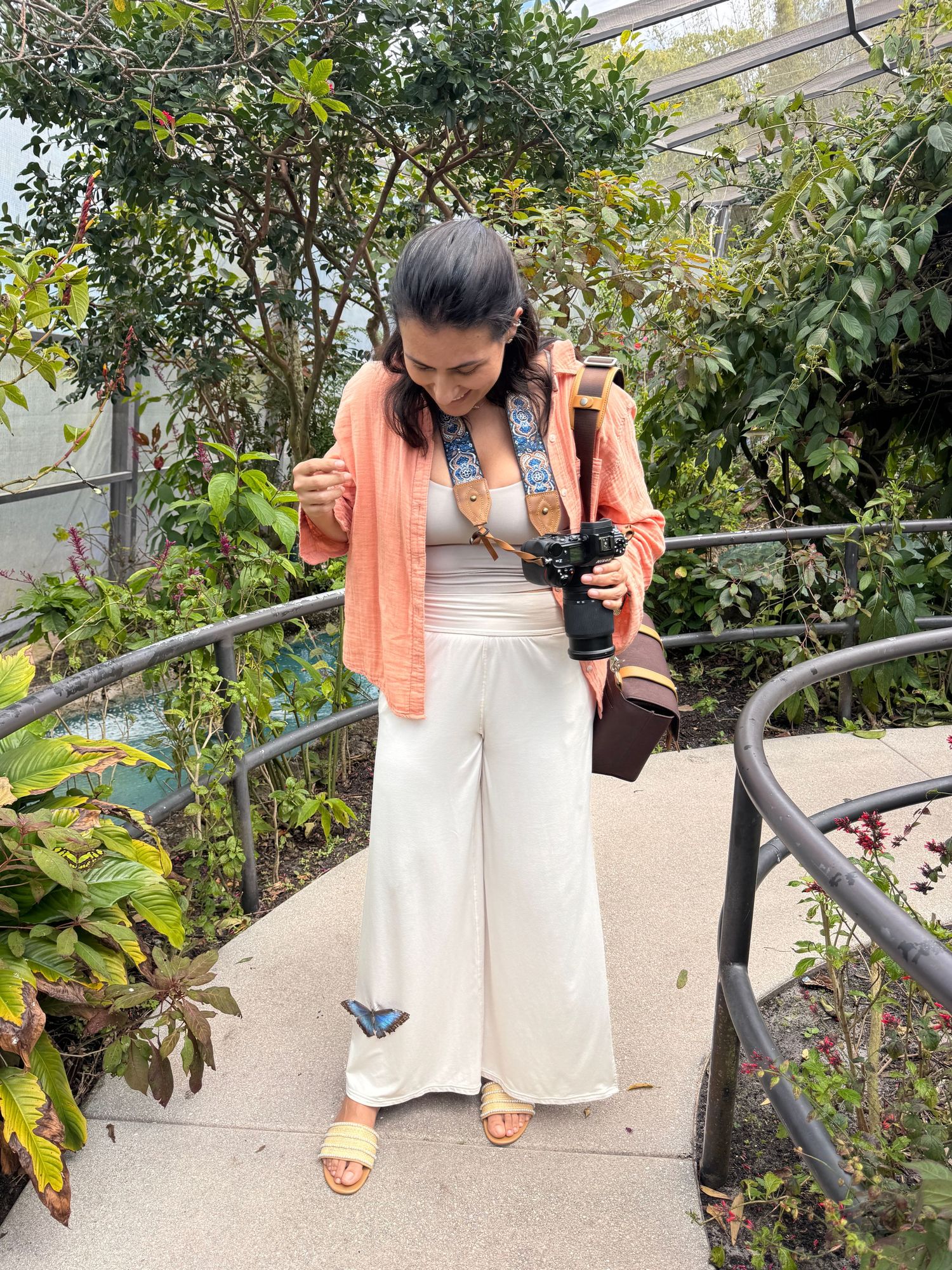 Fashionable outfit featuring white palazzo pants, peach cardigan, and brown accessories in a garden setting.