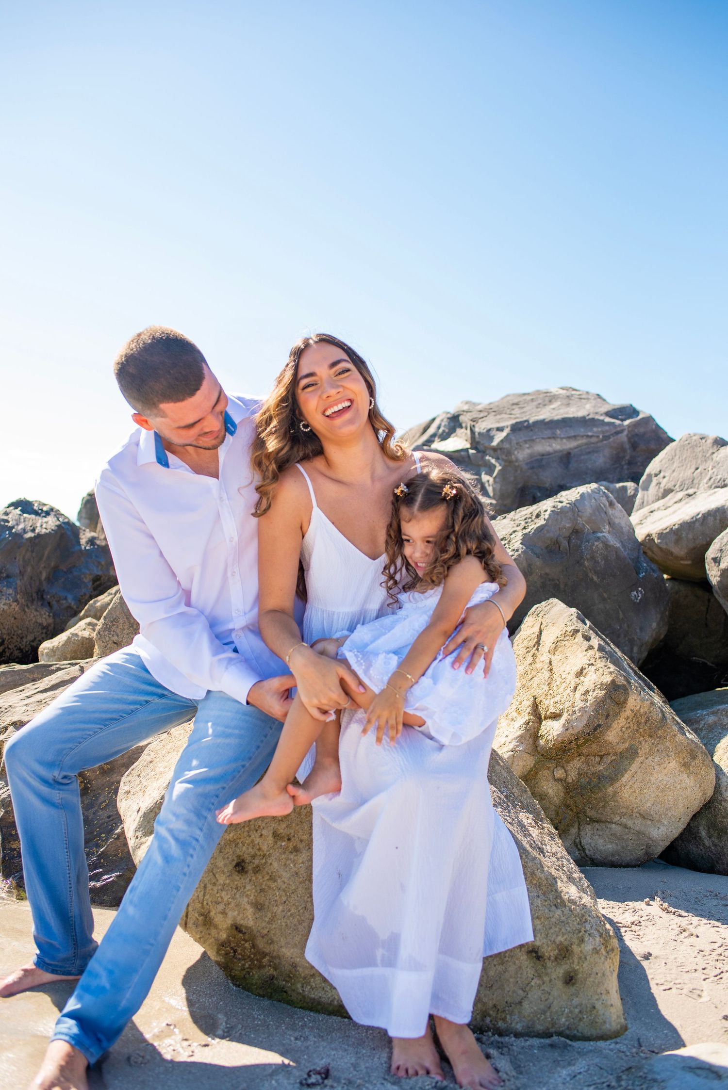Romantic couple in white and denim outfits share intimate moments while posing on rocky beach during sunset photoshoot.