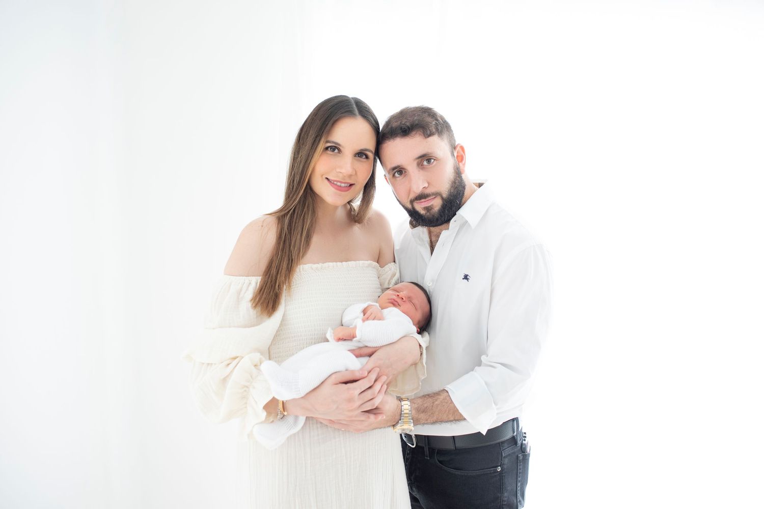 Parents sharing tender moment with newborn baby in all-white studio setting.