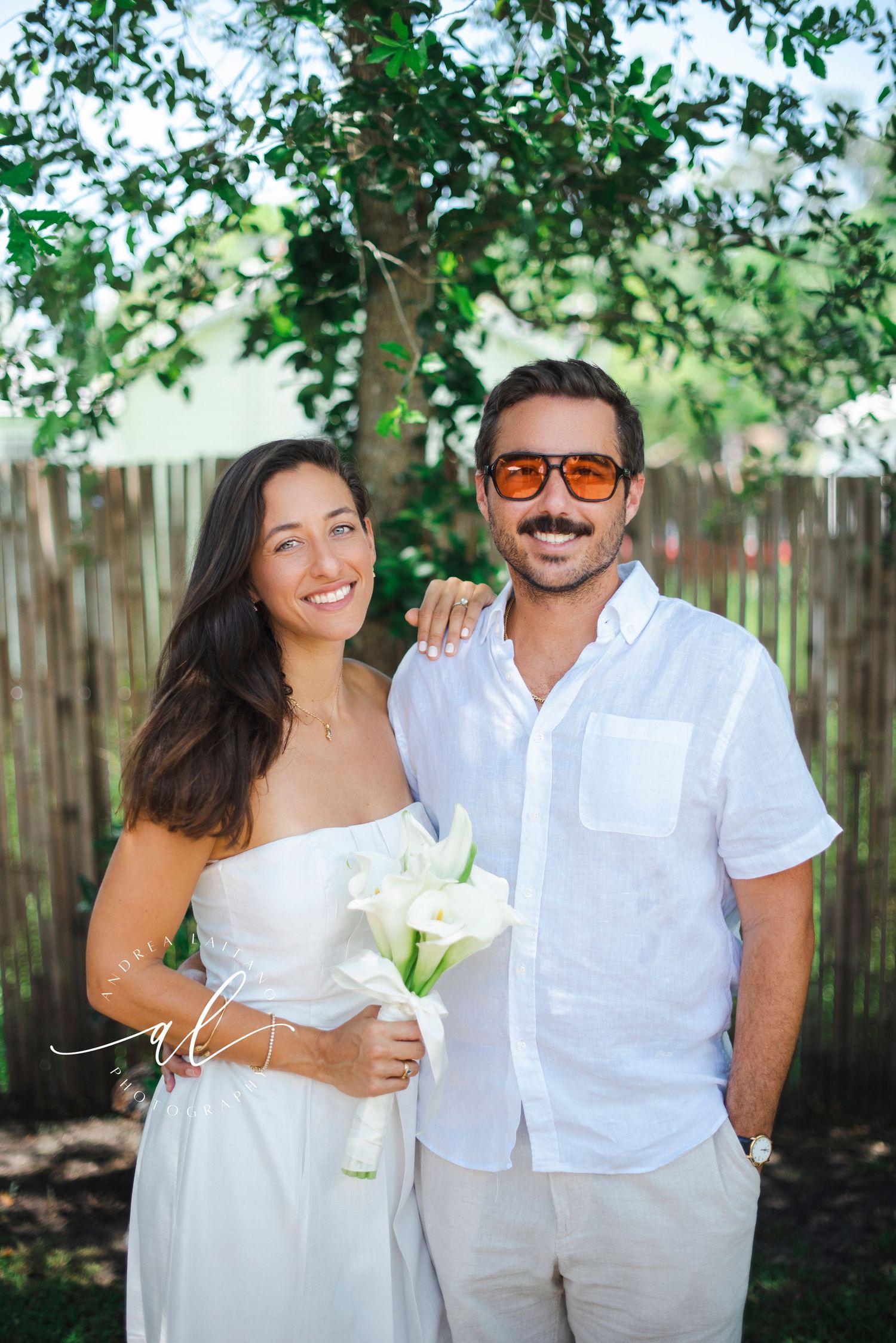 A couple in white outfits posing together in a backyard with green foliage.