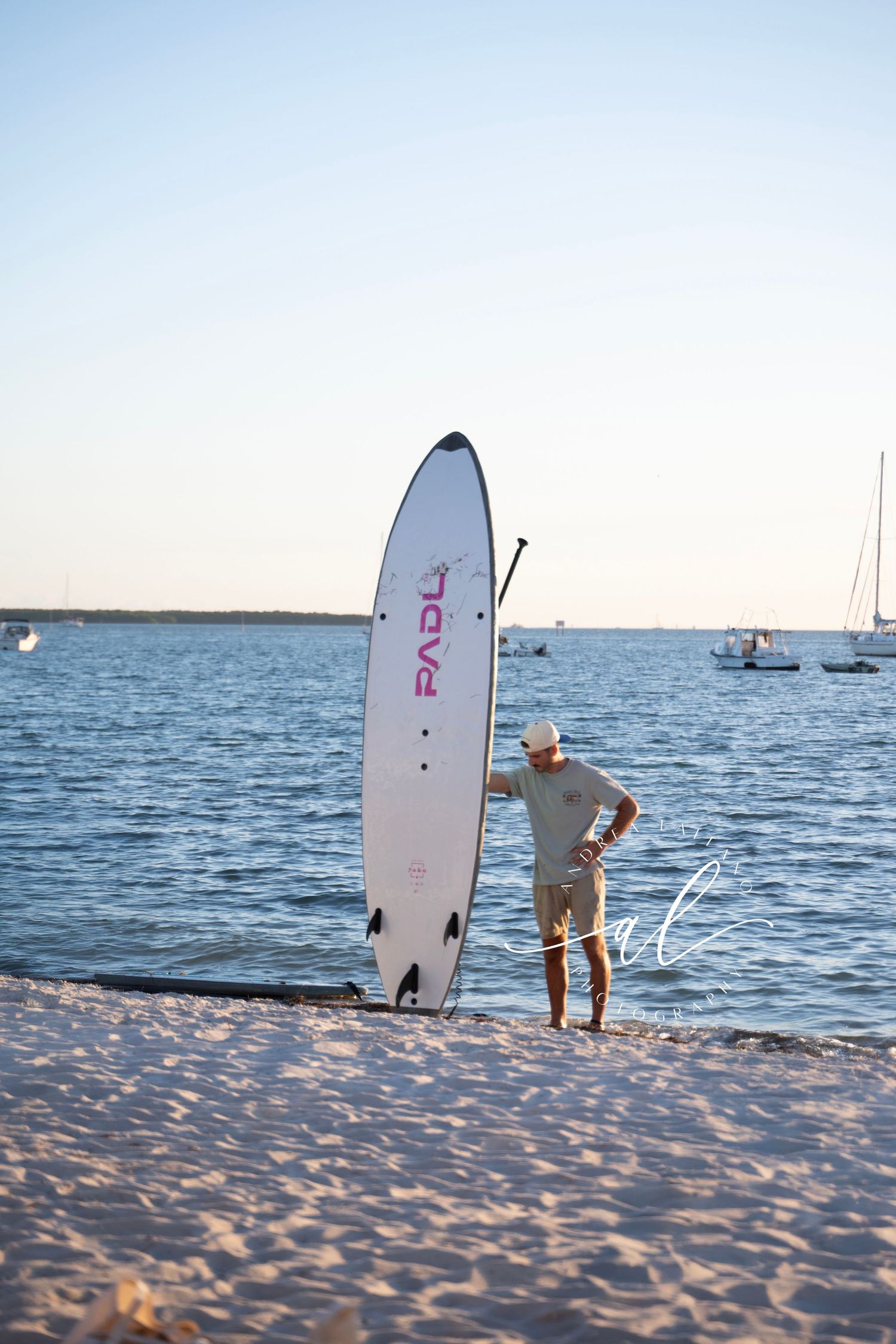 A surfer stands on a sandy beach at sunset with a white surfboard near the ocean.