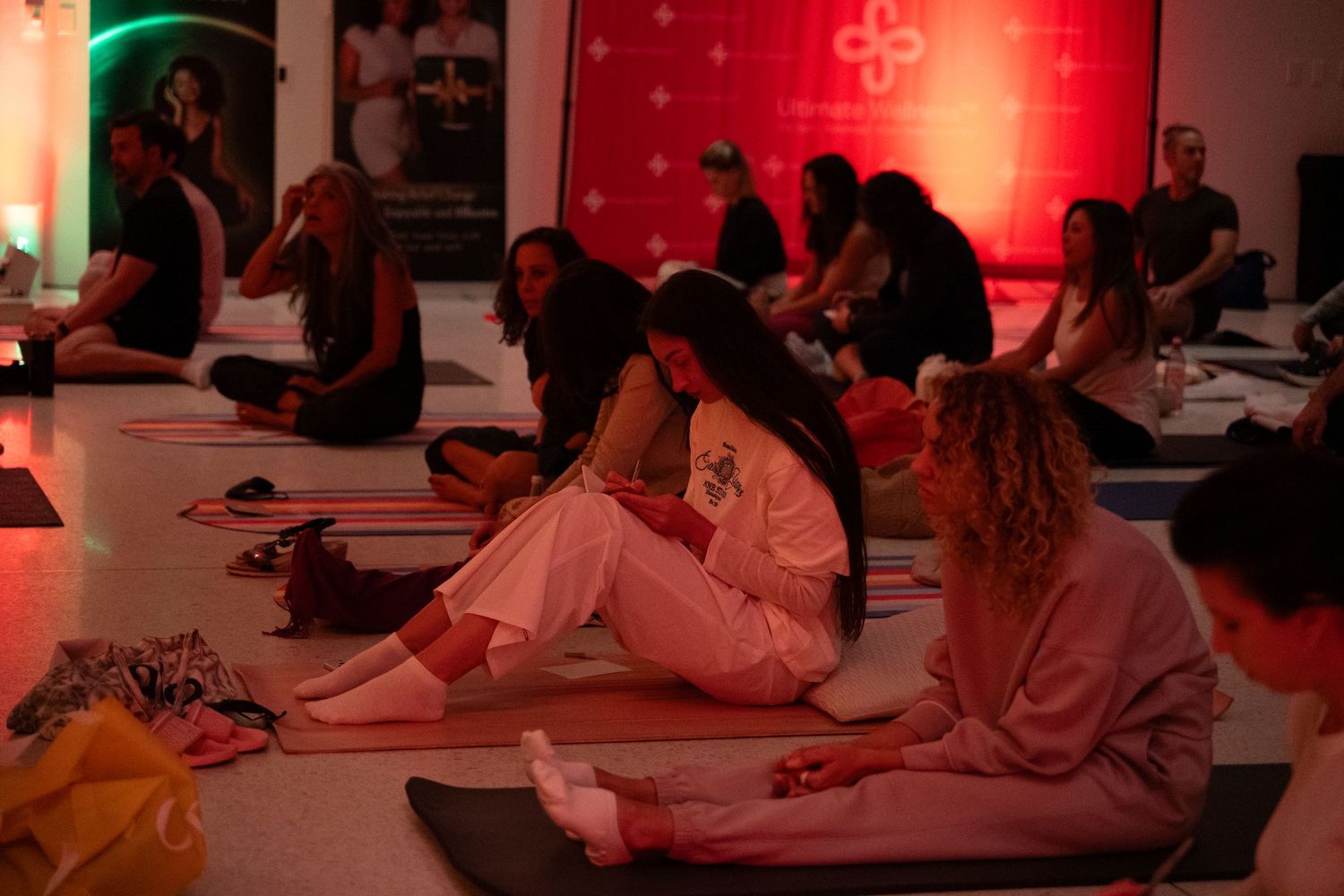 Two women in casual white outfits sit on yoga mats chatting during a yoga class with dramatic red ambient lighting.