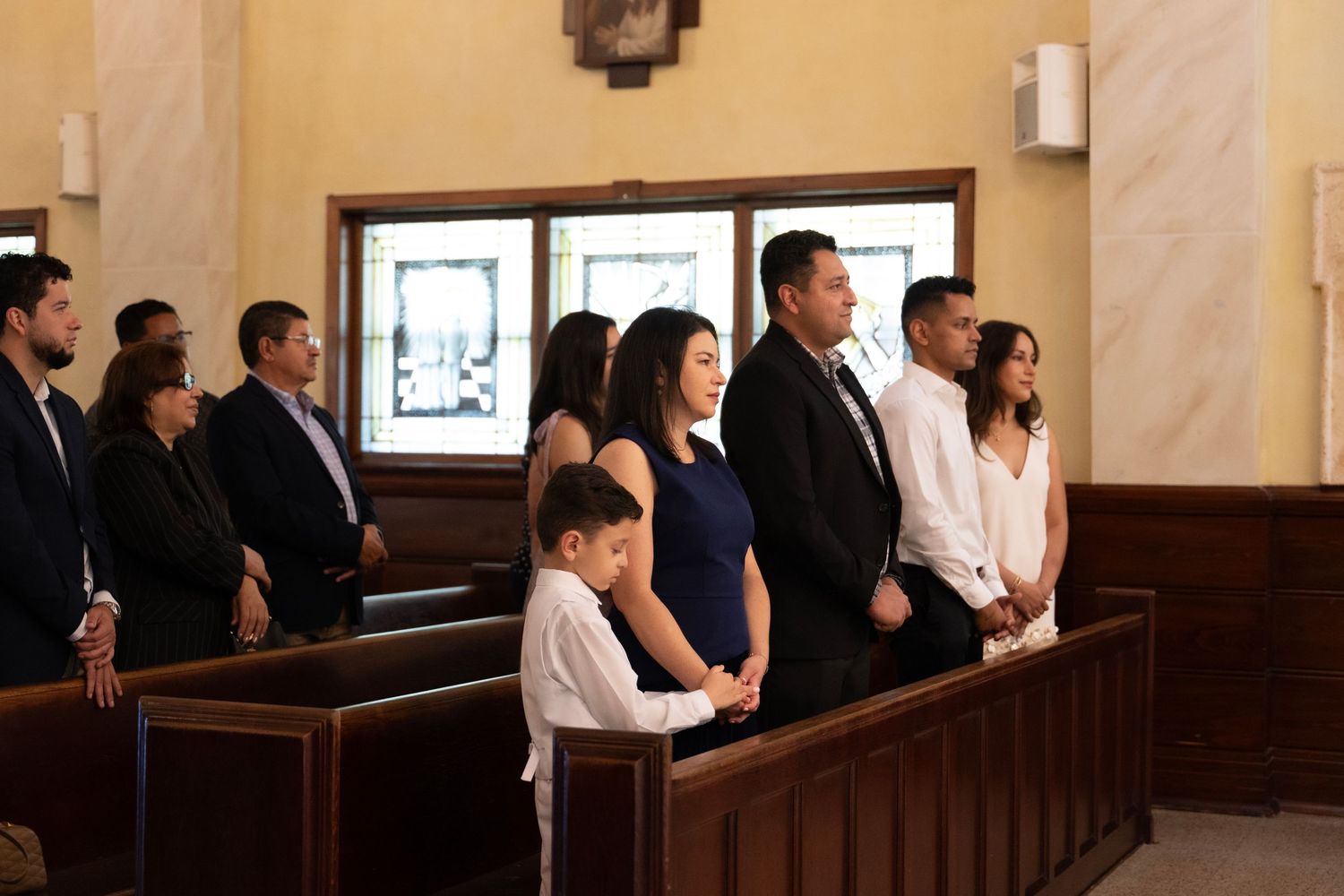 Family standing in church pews during a religious ceremony, dressed in formal attire.