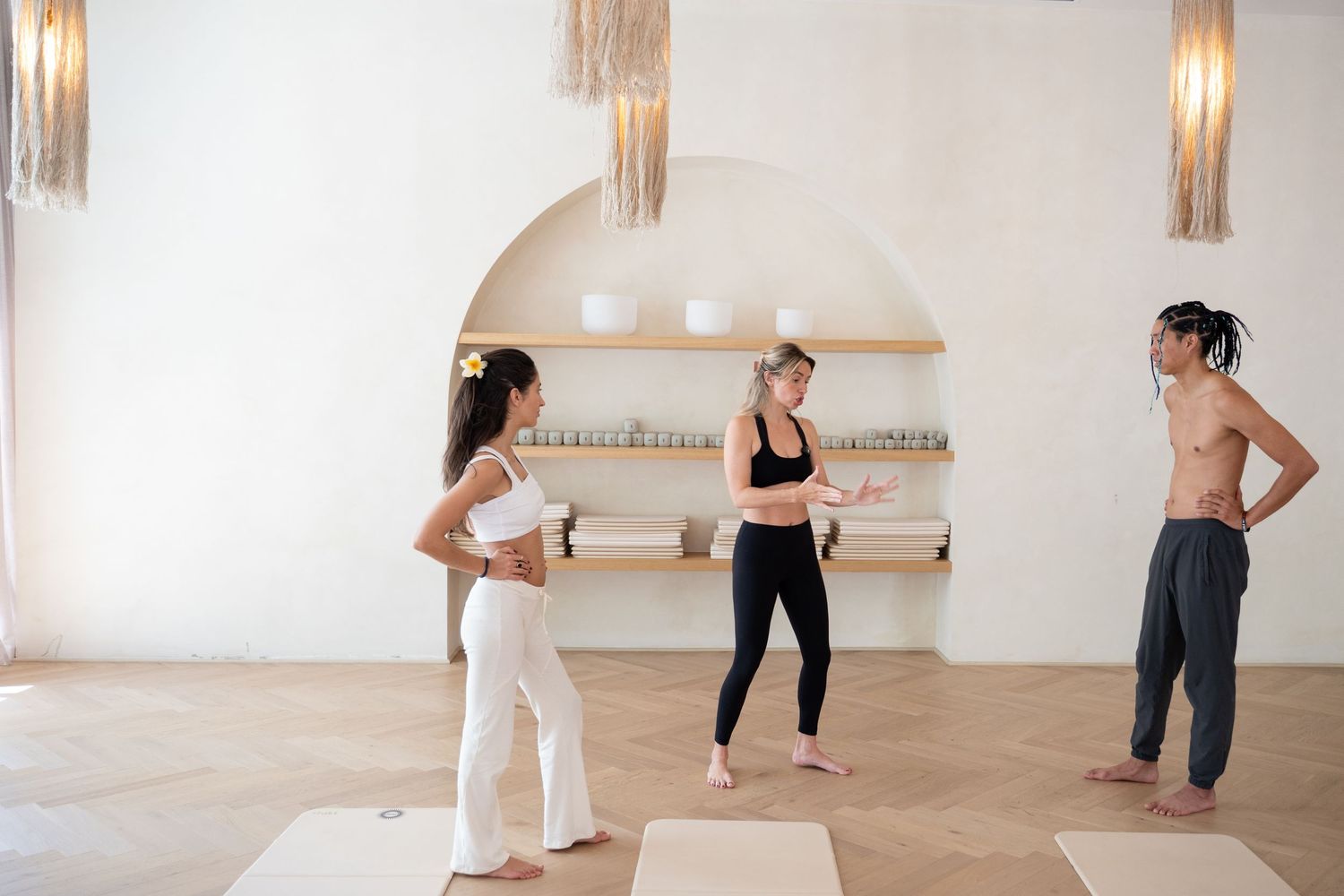 Three people standing on yoga mats in a bright, modern studio with shelves and pendant lights.