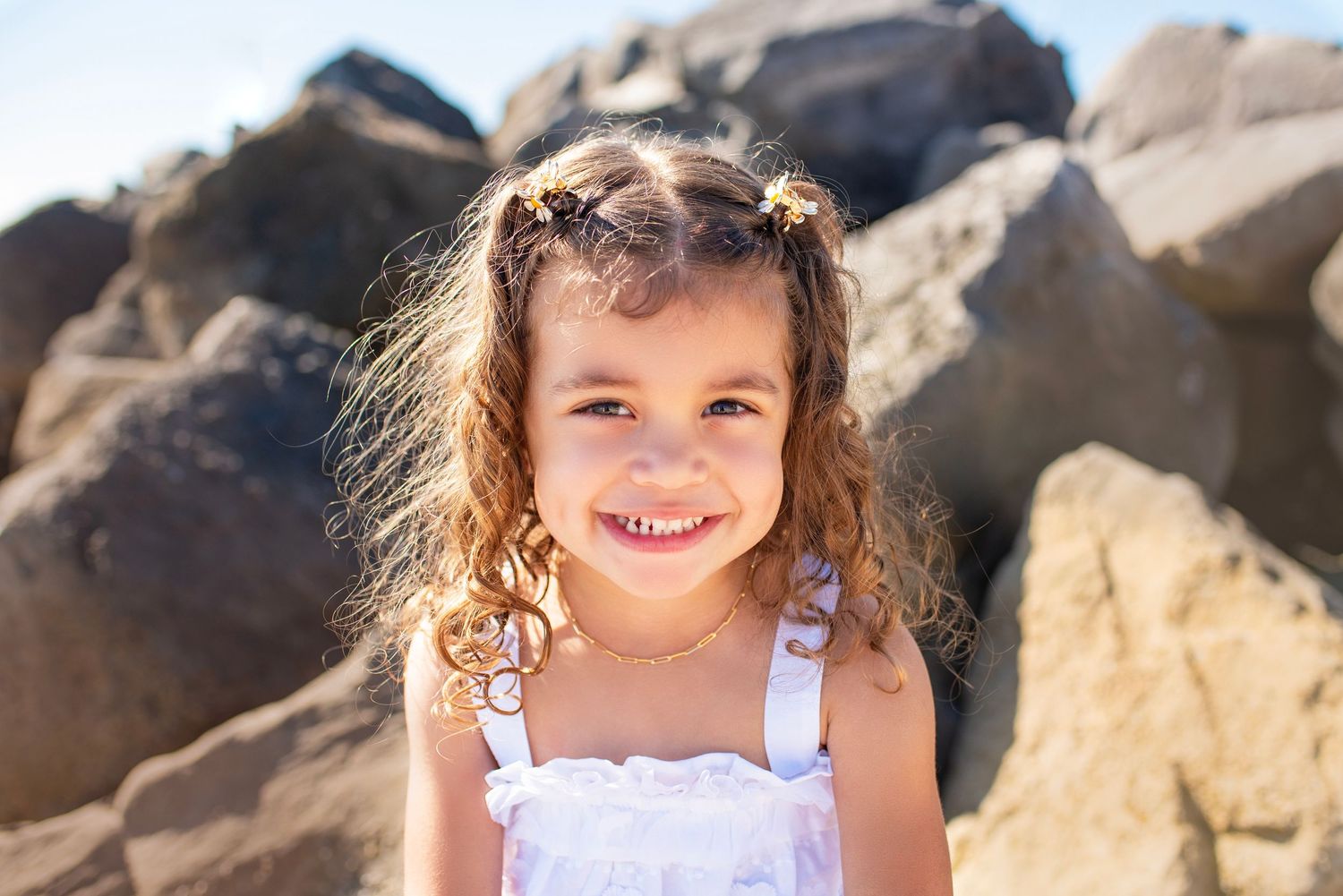 Young child with curly hair wearing a white dress smiles brightly against a backdrop of large rocks on a sunny day.