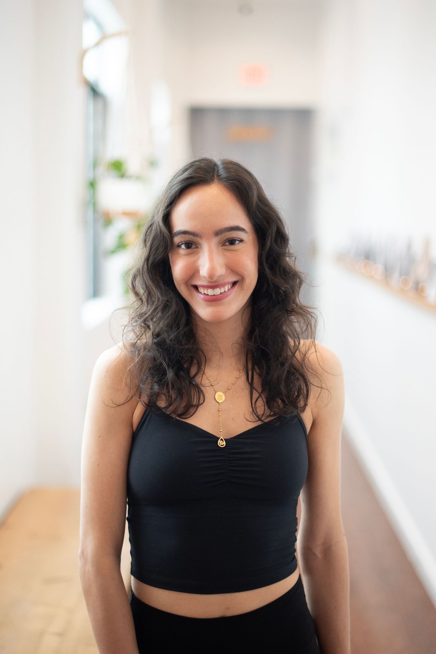 A person with dark curly hair wearing a black crop top smiles in a bright indoor setting.