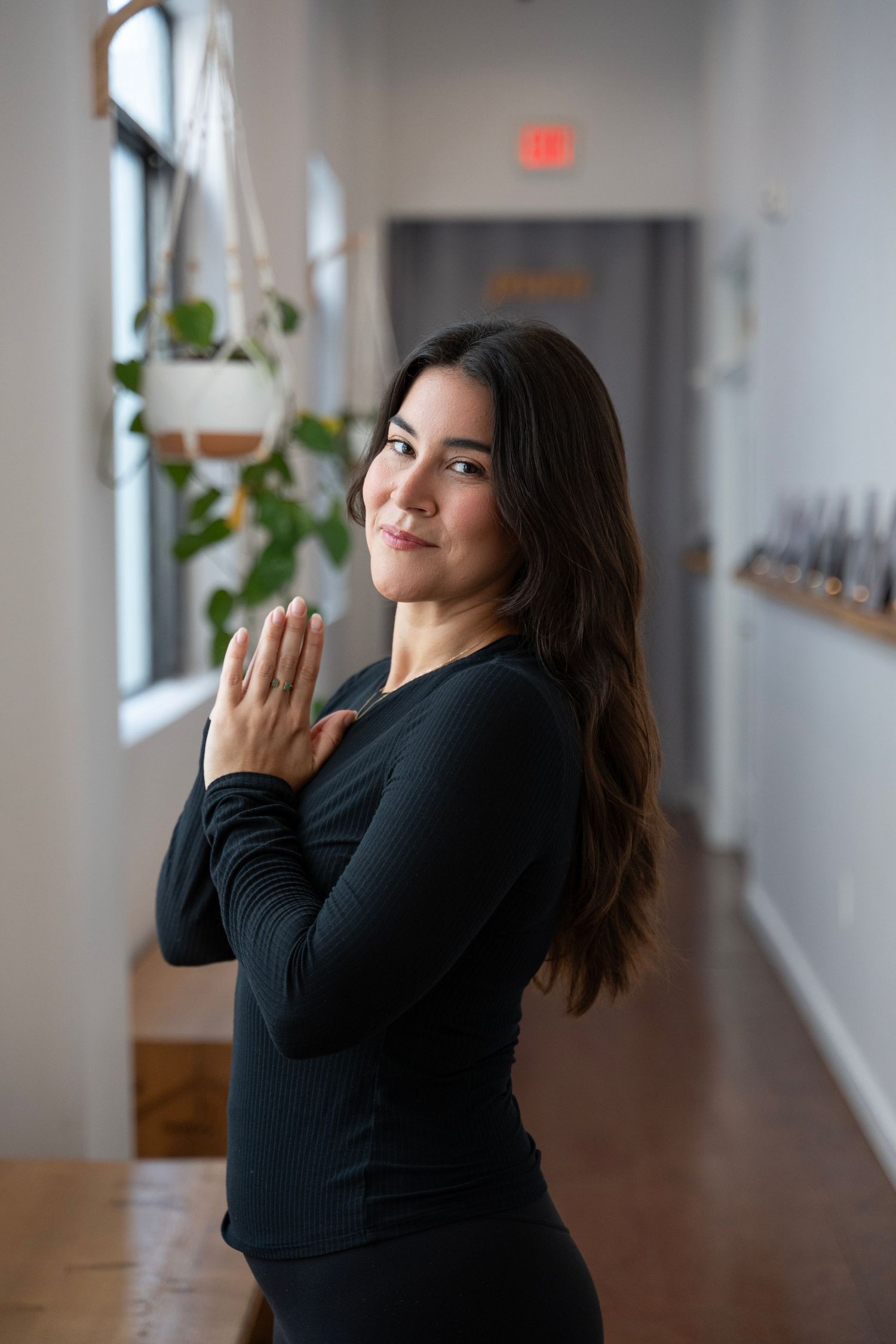 Person in black long-sleeved top poses thoughtfully in a bright hallway with hanging plants and natural light.