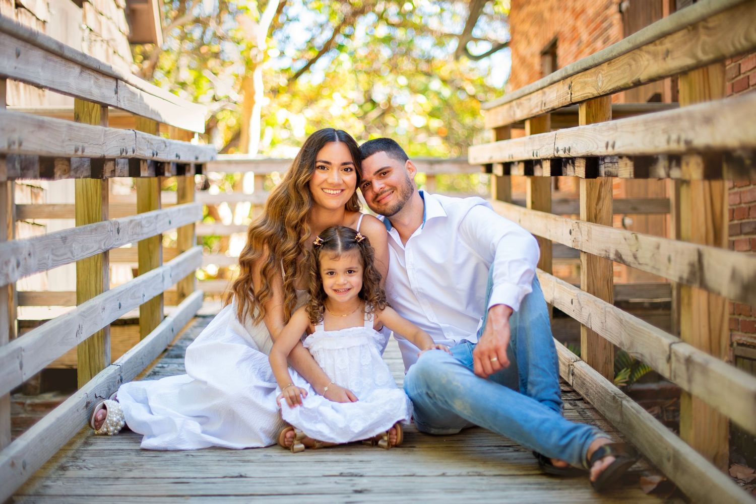 A family wearing white sits together on a wooden bridge during a sunny outdoor photo session.