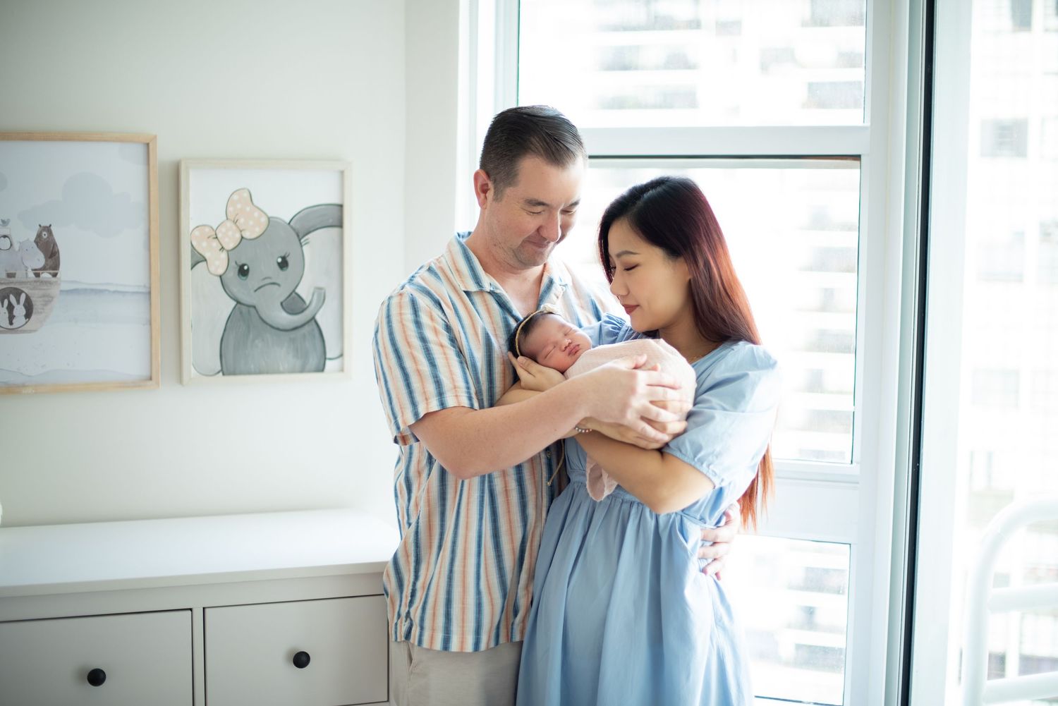 Parents in soft blue and striped clothing hold their newborn baby near a nursery window with framed artwork.