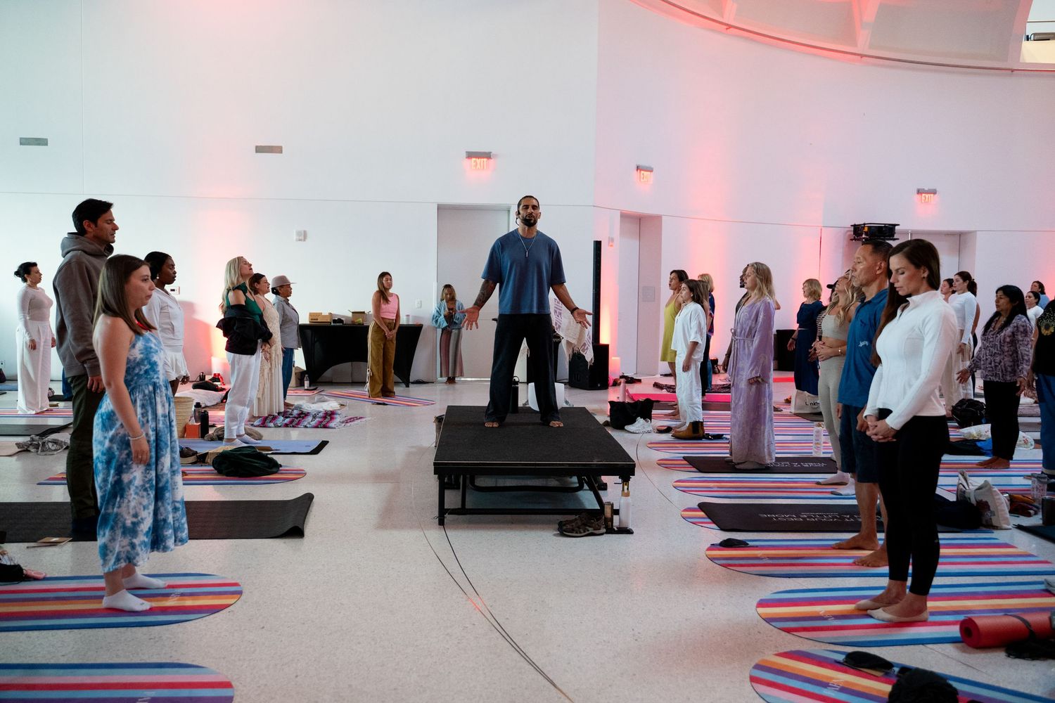 A yoga instructor leads a large group class on colorful striped mats in a bright white studio with warm pink lighting.