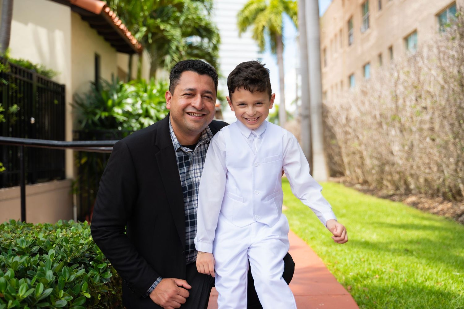 A man and young boy in white outfit smiling together outdoors on a brick path with green lawn and palm trees.