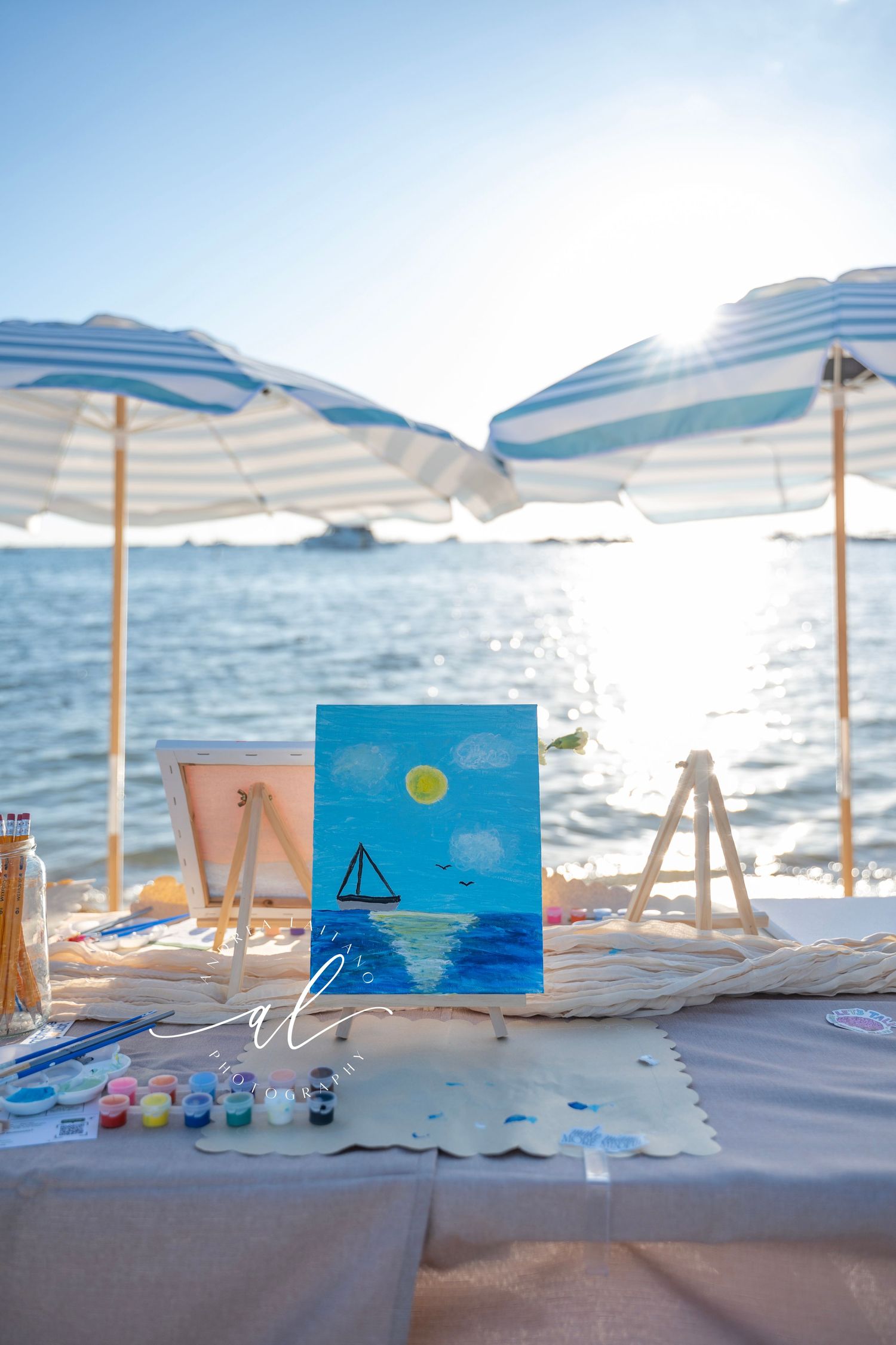 Beach art setup with easels, umbrellas, and painting supplies along a serene oceanfront on a sunny day.