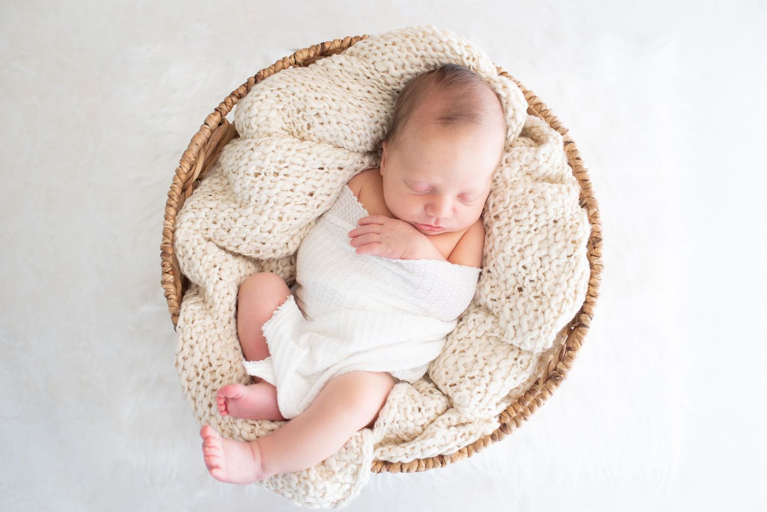 Peaceful newborn baby resting in woven basket with cream knit blanket.