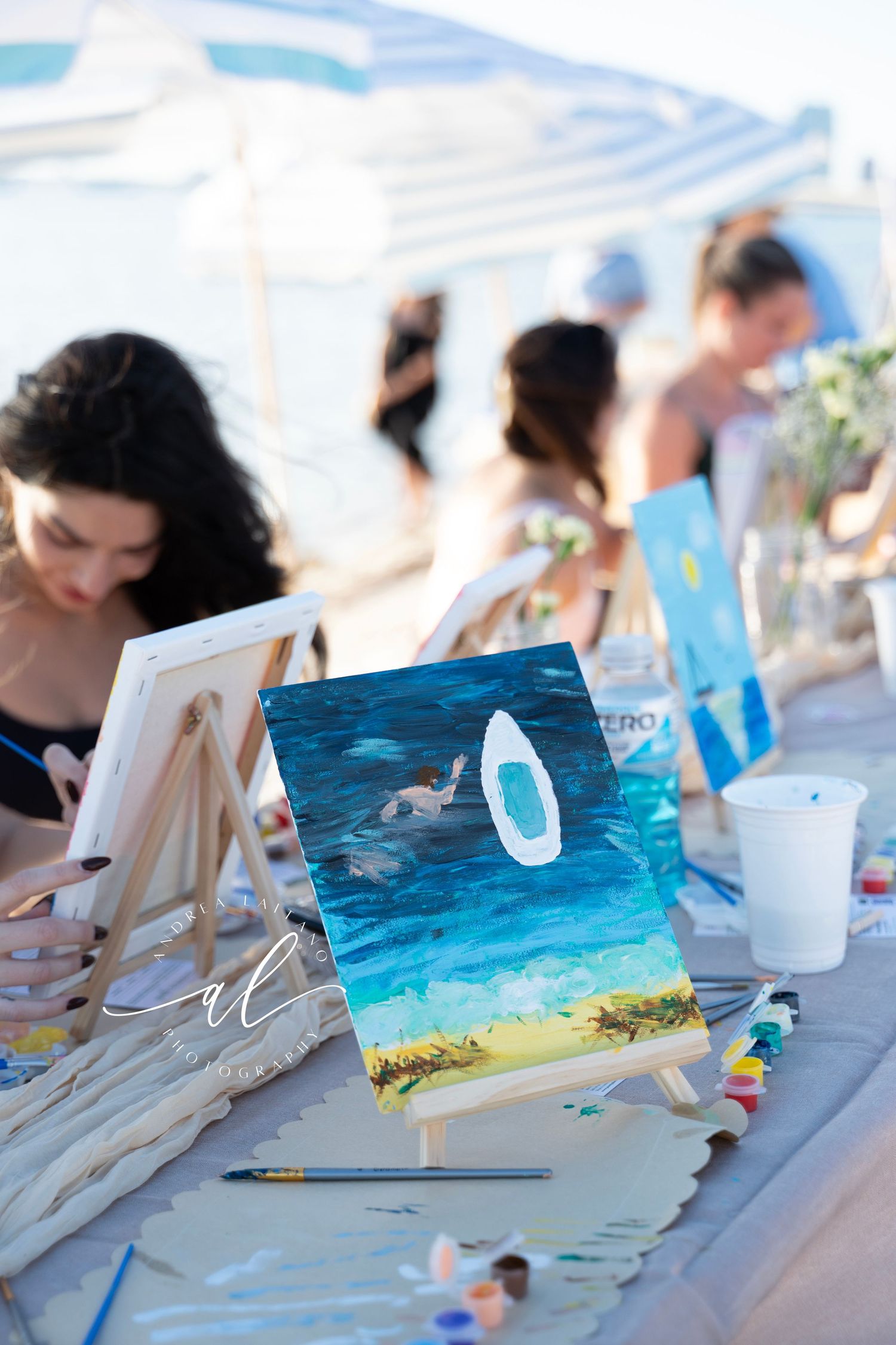 A small painting on an easel shows a whale tail emerging from blue ocean waters during an outdoor art class session.