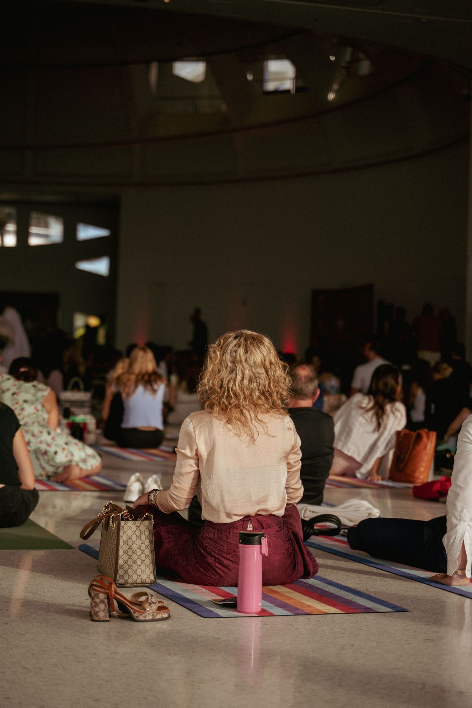Woman with curly hair sits on yoga mat in crowded indoor group meditation session with pink water bottle nearby.