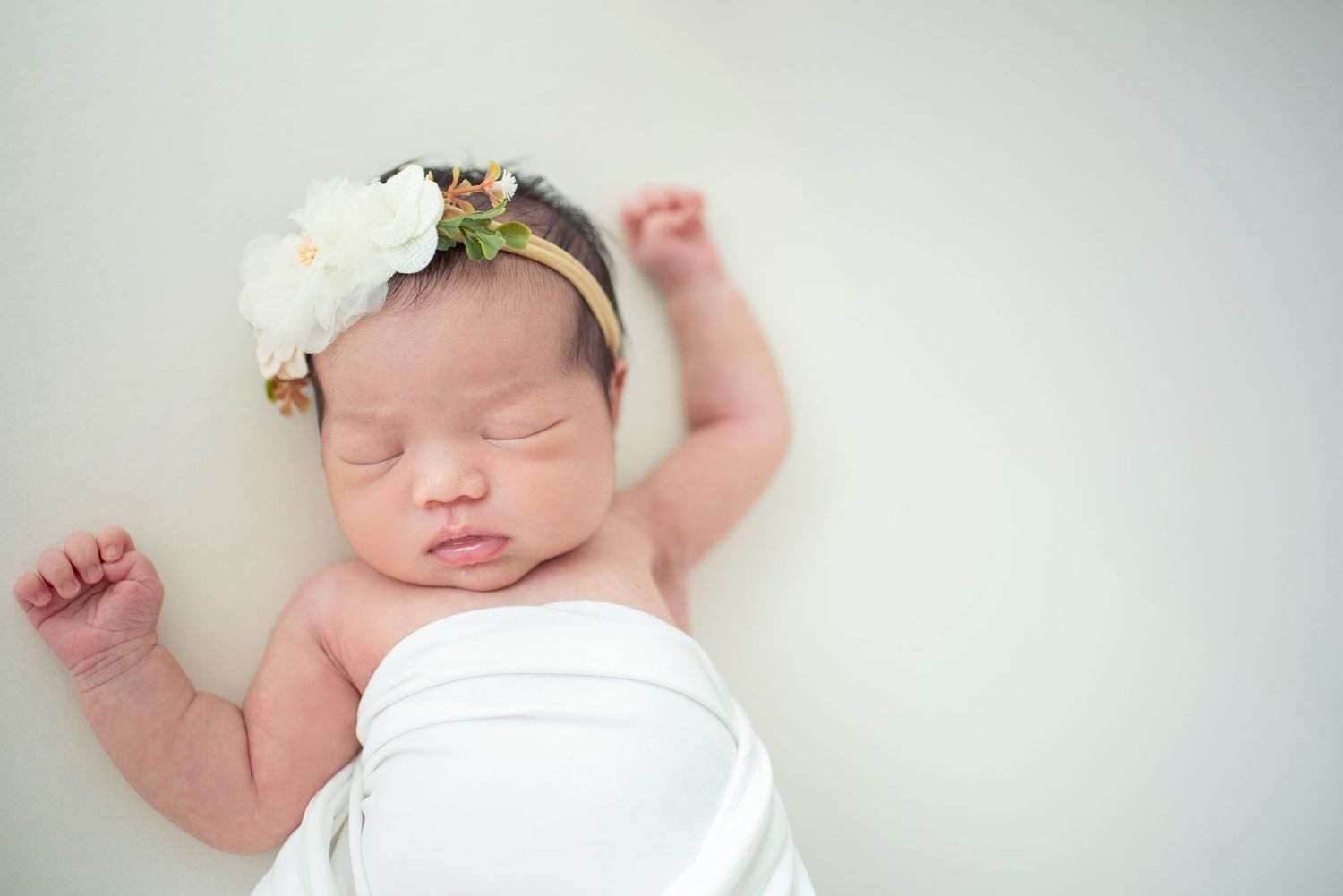 A sleeping newborn baby wrapped in soft white fabric lies peacefully against a light background.