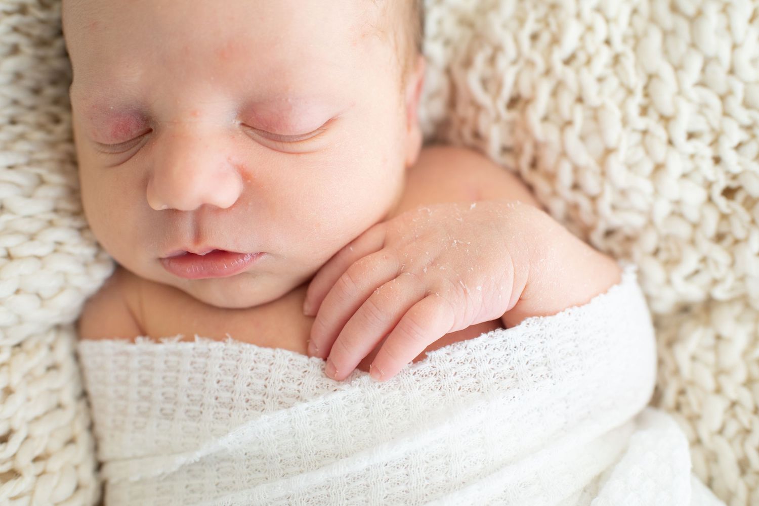 Delicate close-up details of a sleeping newborn wrapped in soft white textiles against a cream background.